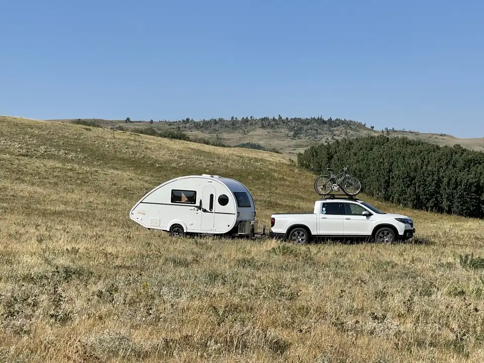 White pickup truck towing a small white camper across a dry grassy hillside under a blue sky