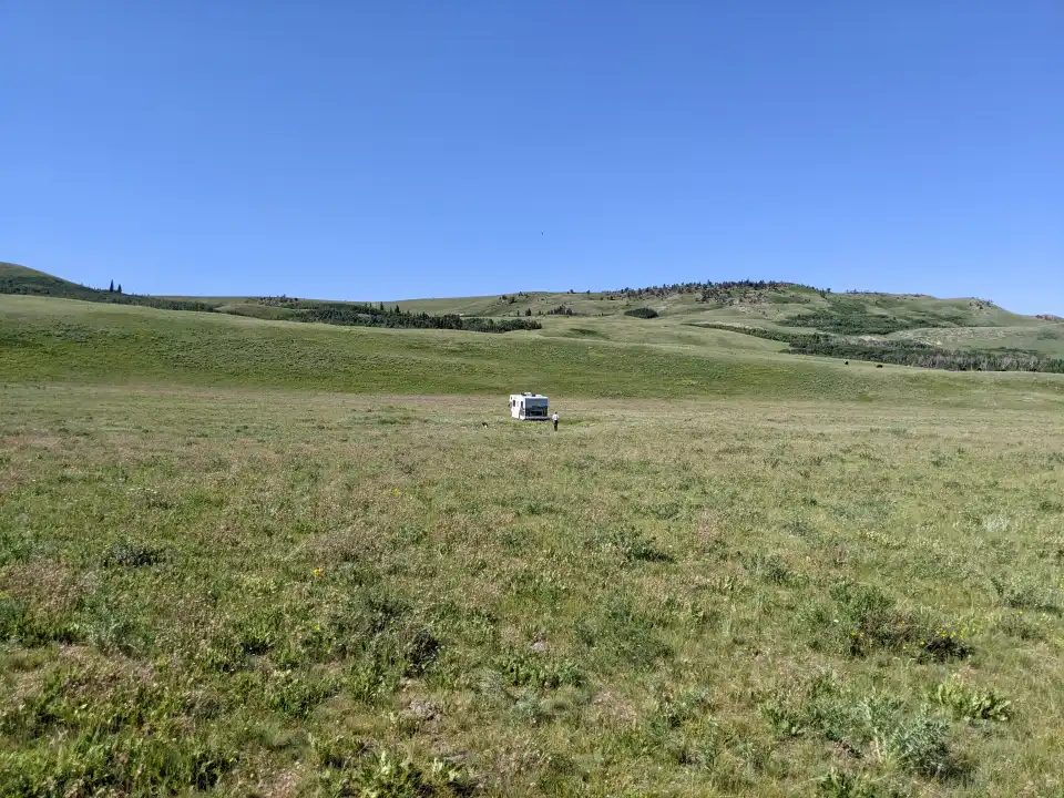Small white car in a grassy field under a clear blue sky with rolling green hills.
