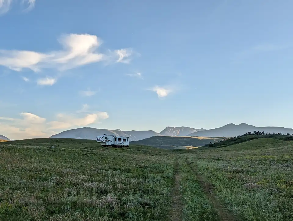 Grassy field with a dirt track leading to a white house, mountains in the distance under a blue sky.