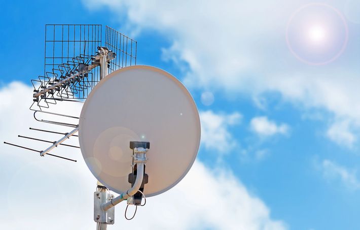 Parabola satellitare e antenna TV sul tetto contro un cielo azzurro con nuvole e bagliori di sole.