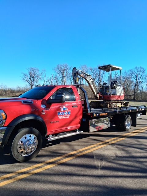 Red tow truck with small excavator on a flatbed, parked on a road, with a blue sky in the background.