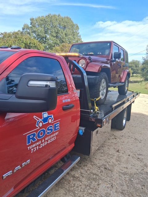 Red tow truck hauling a red Jeep Wrangler on a flatbed. Outdoors, sunny day.