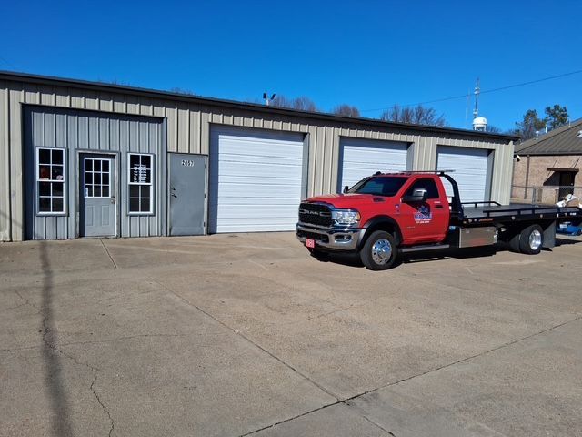 Red tow truck parked in front of a metal building with three garage doors; sunny day.