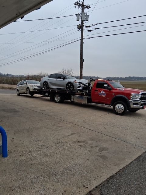 Red tow truck carrying two cars on a flatbed trailer, in an overcast outdoor setting.