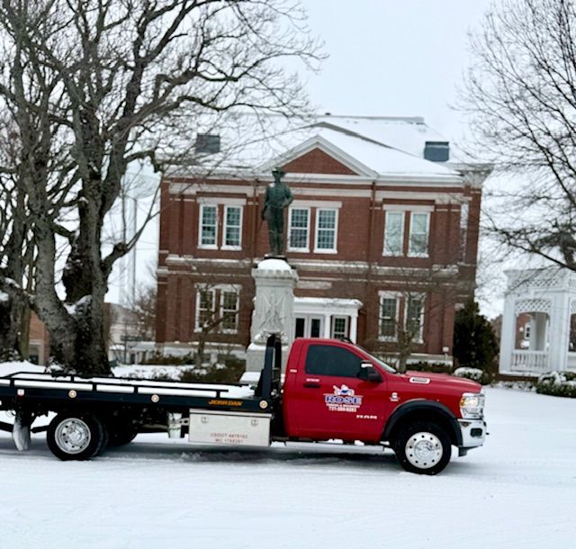 Red tow truck in snowy setting in front of a brick building and a statue.