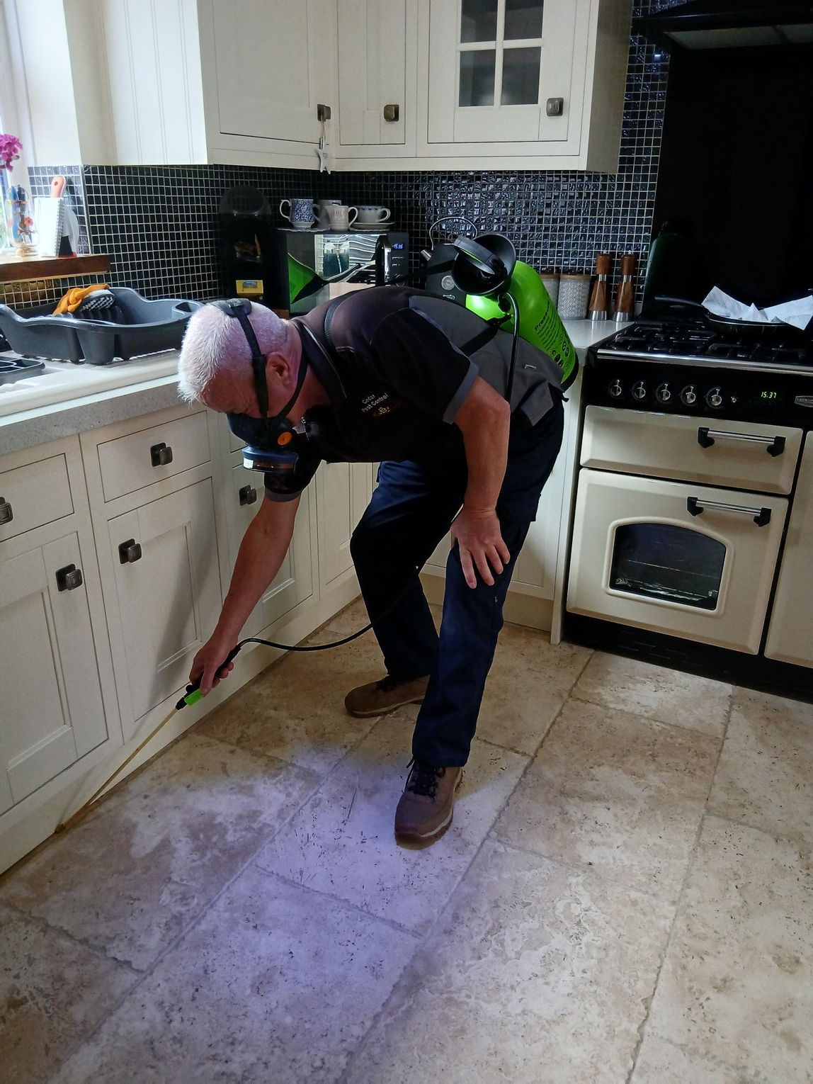 Person kneeling in a kitchen, cleaning the floor with a broom near white cabinets and a stove.