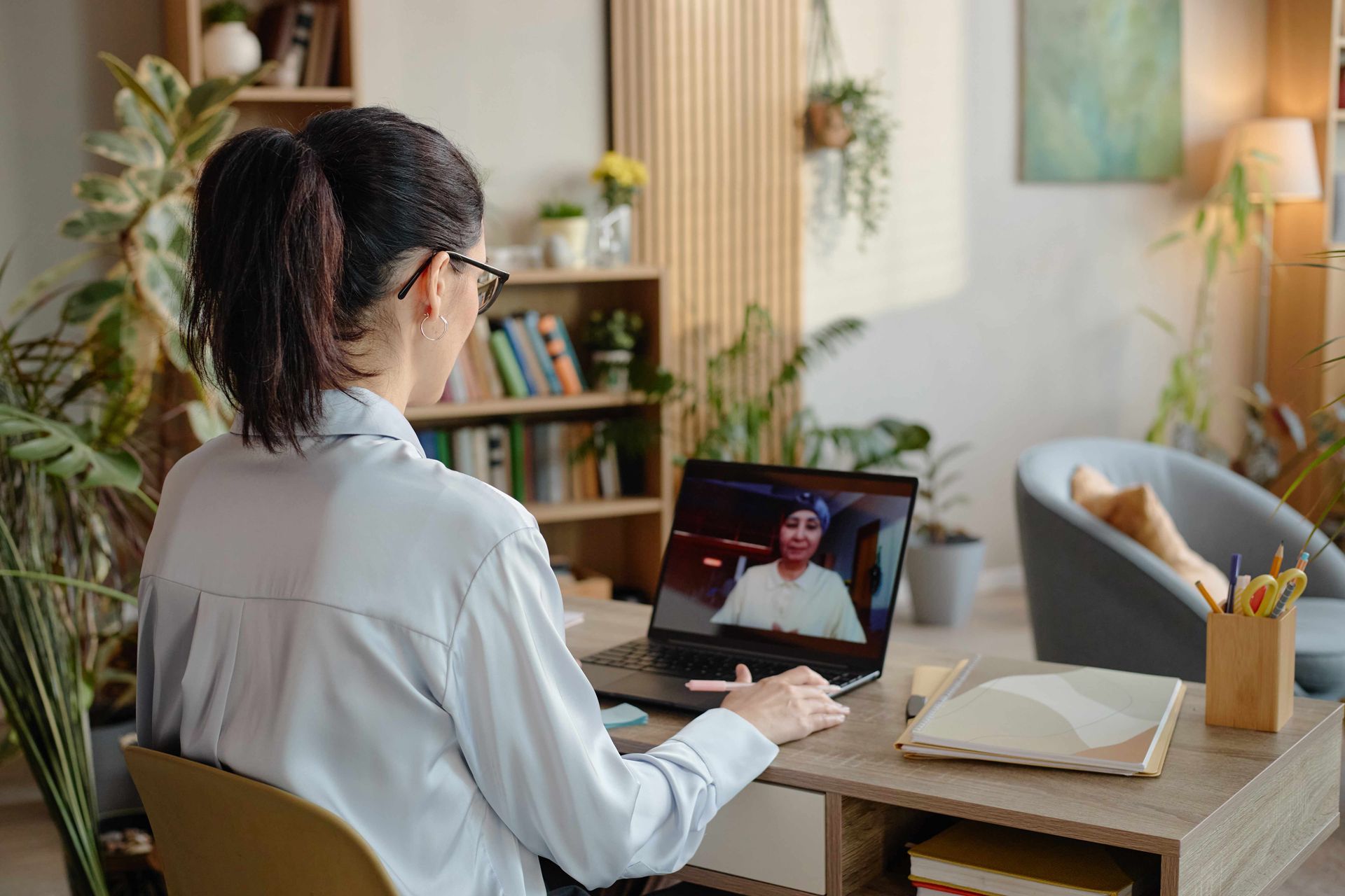 Person working on a laptop at a dining table during a video call, with books and a lamp nearby Person working on a laptop at a dining table during a video call, with books and a lamp nearby