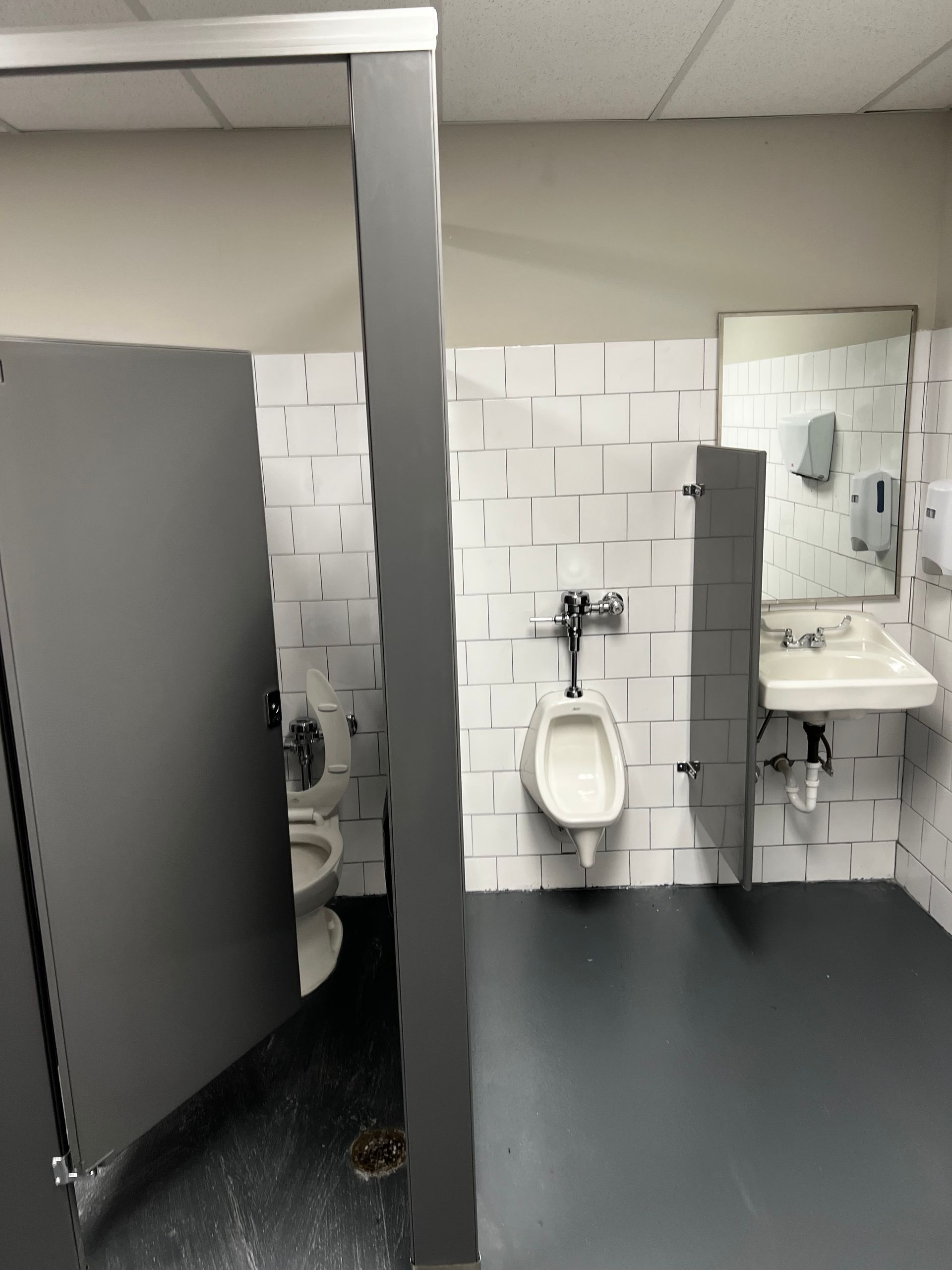 Public restroom with a toilet stall, wall-mounted urinal, sink, mirrors, and gray flooring.