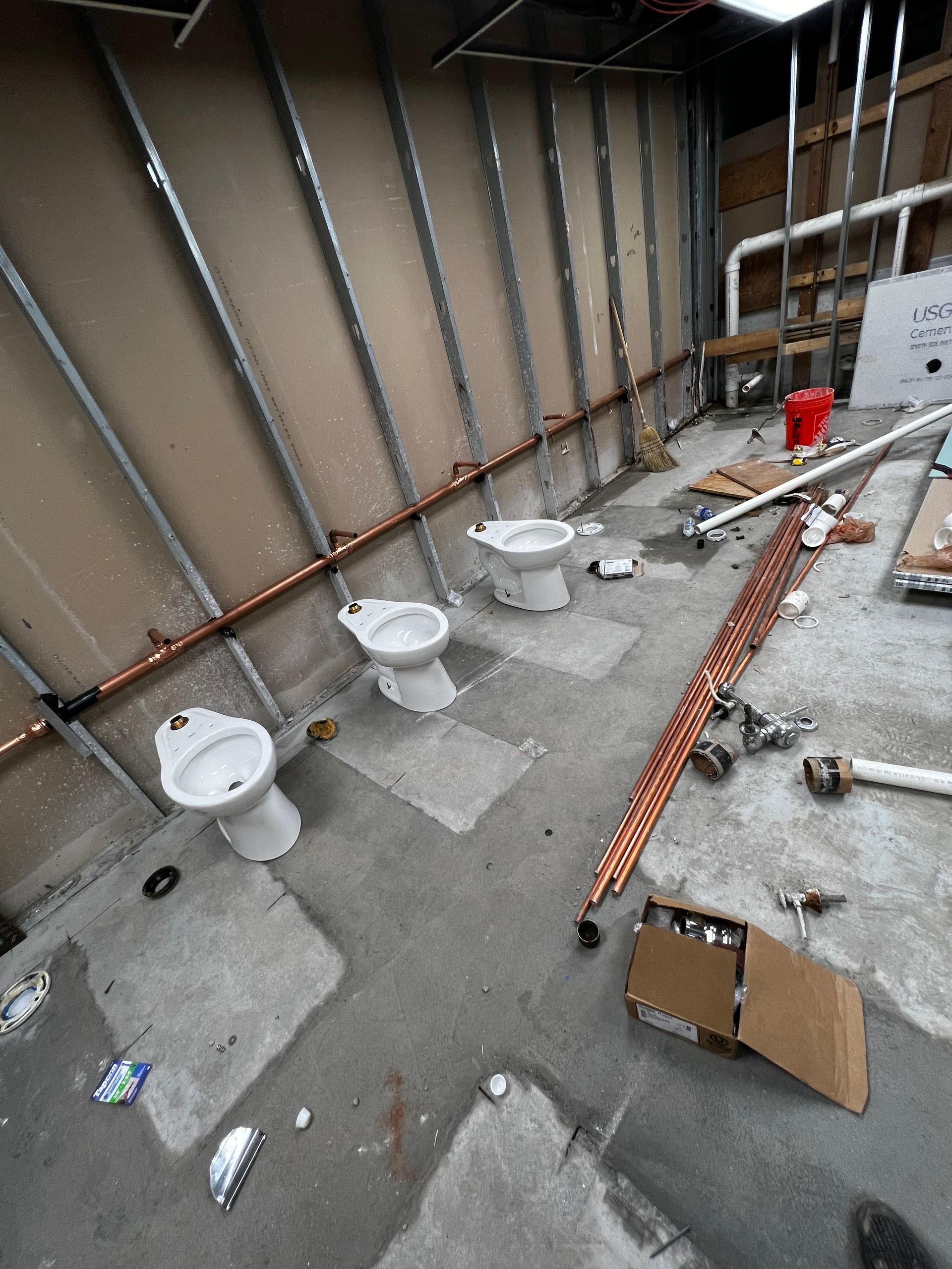 Construction site with three toilets, scattered tools, and orange conduit against a wall