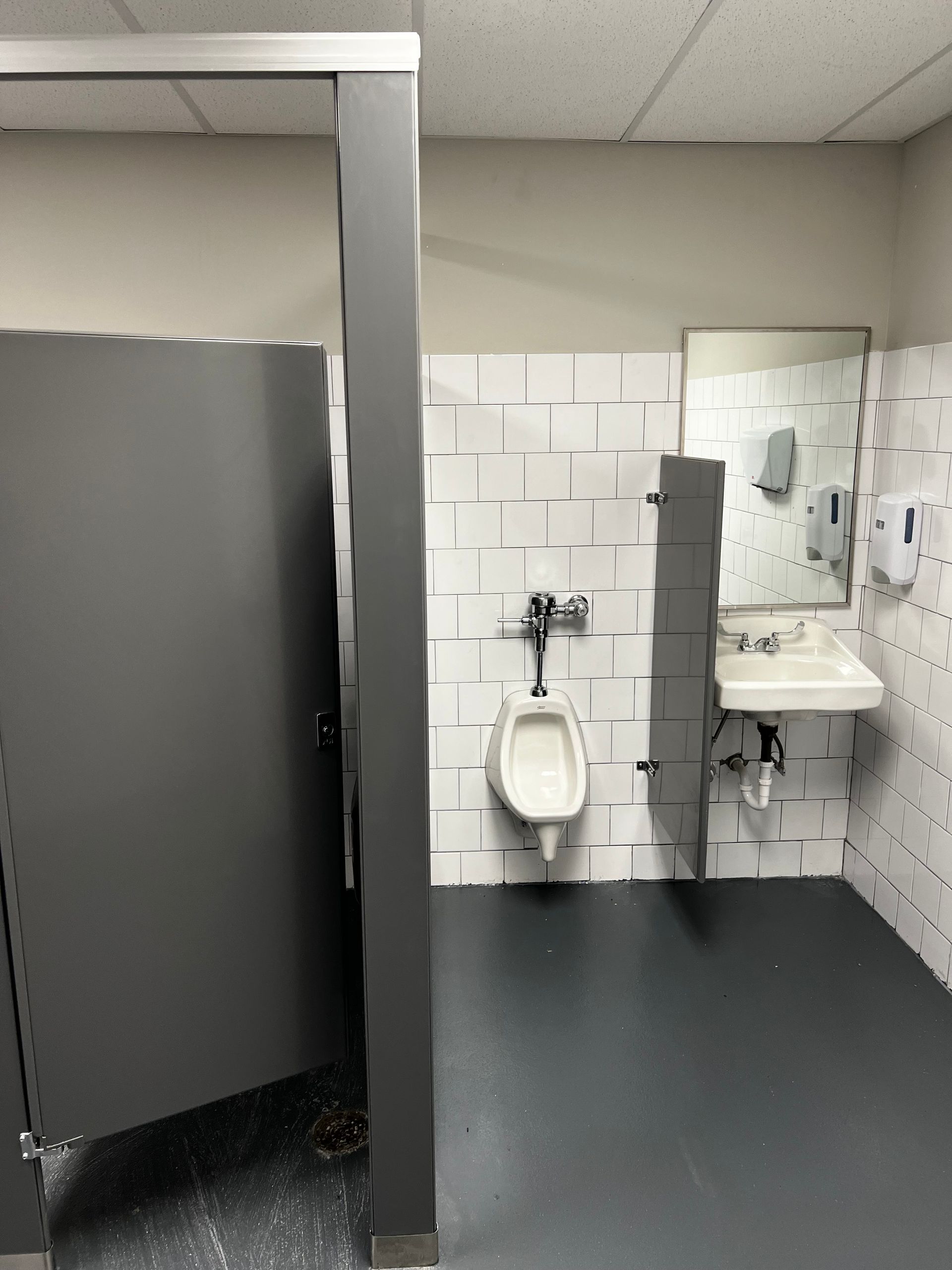 Public restroom with a urinal, sink, mirror, and gray stall partitions on a tiled wall.