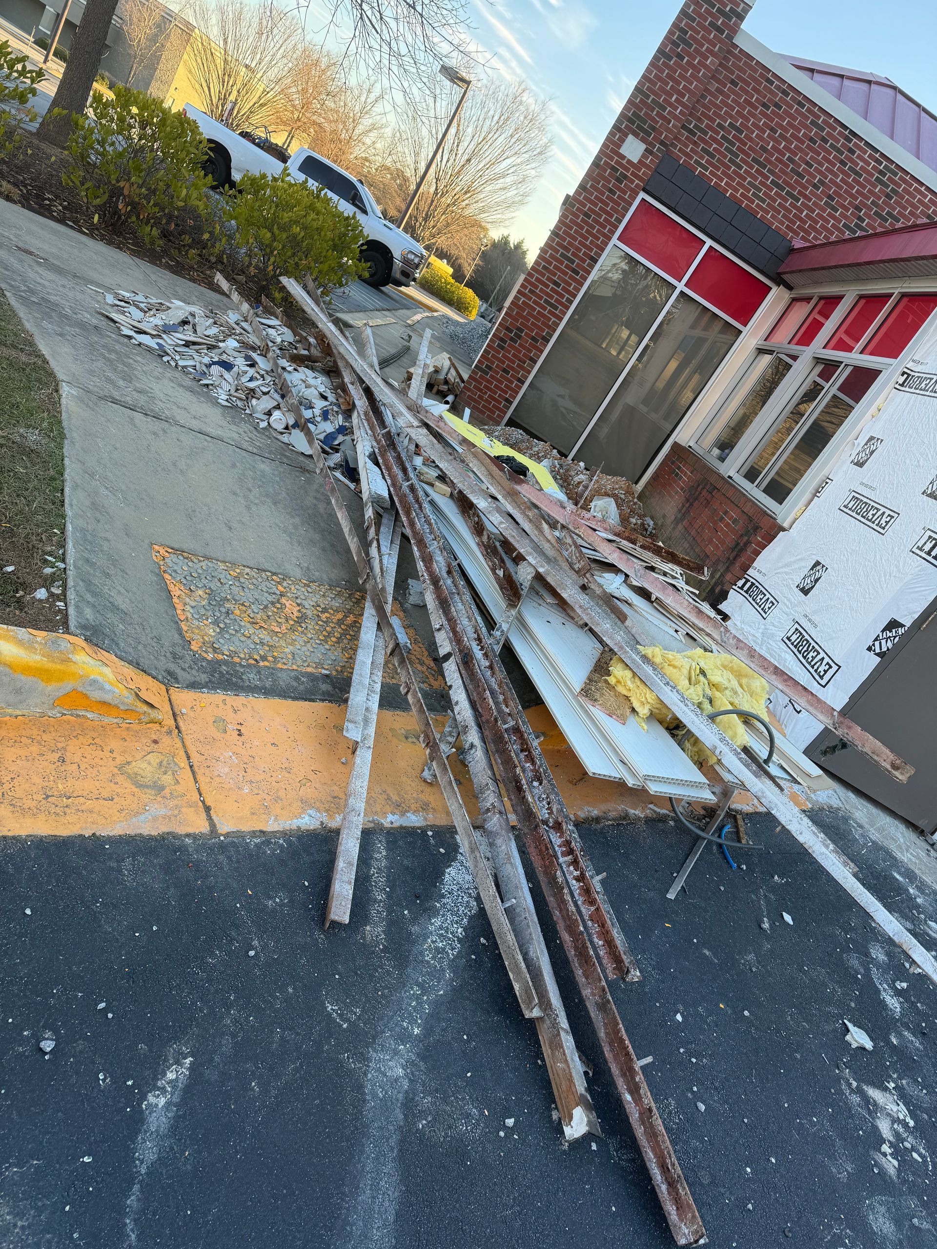 Pile of long metal poles on a sidewalk beside a brick building and red-trimmed windows
