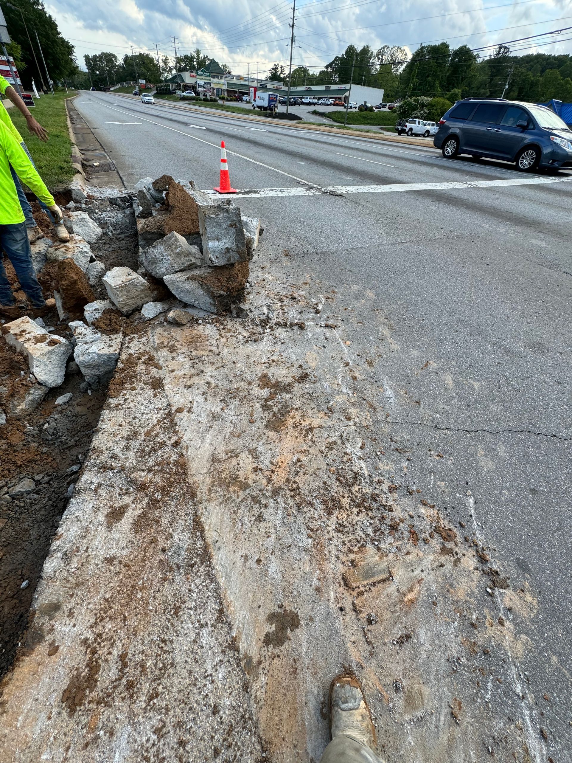 Damaged roadside shoulder with large rocks, gravel, and a traffic cone beside a wet road under repair