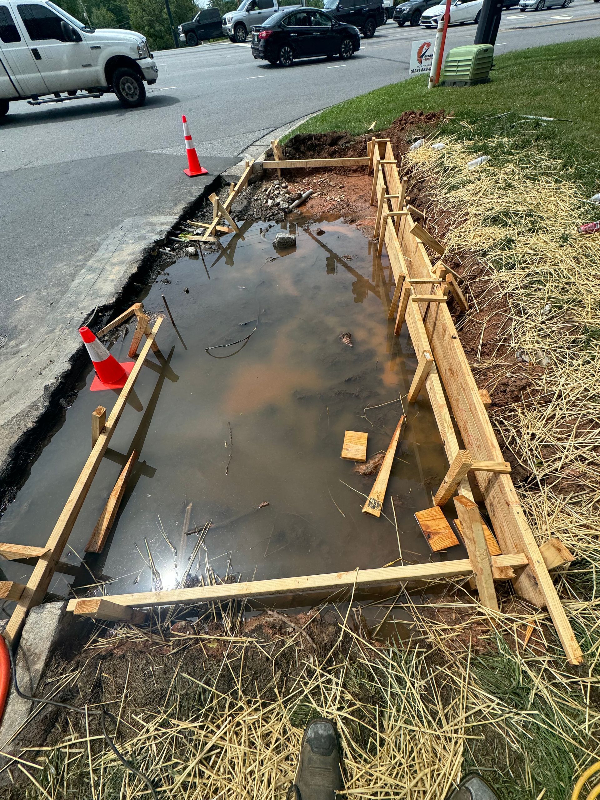 Curbside utility trench filled with water, lined with wood forms and orange cones beside a street.