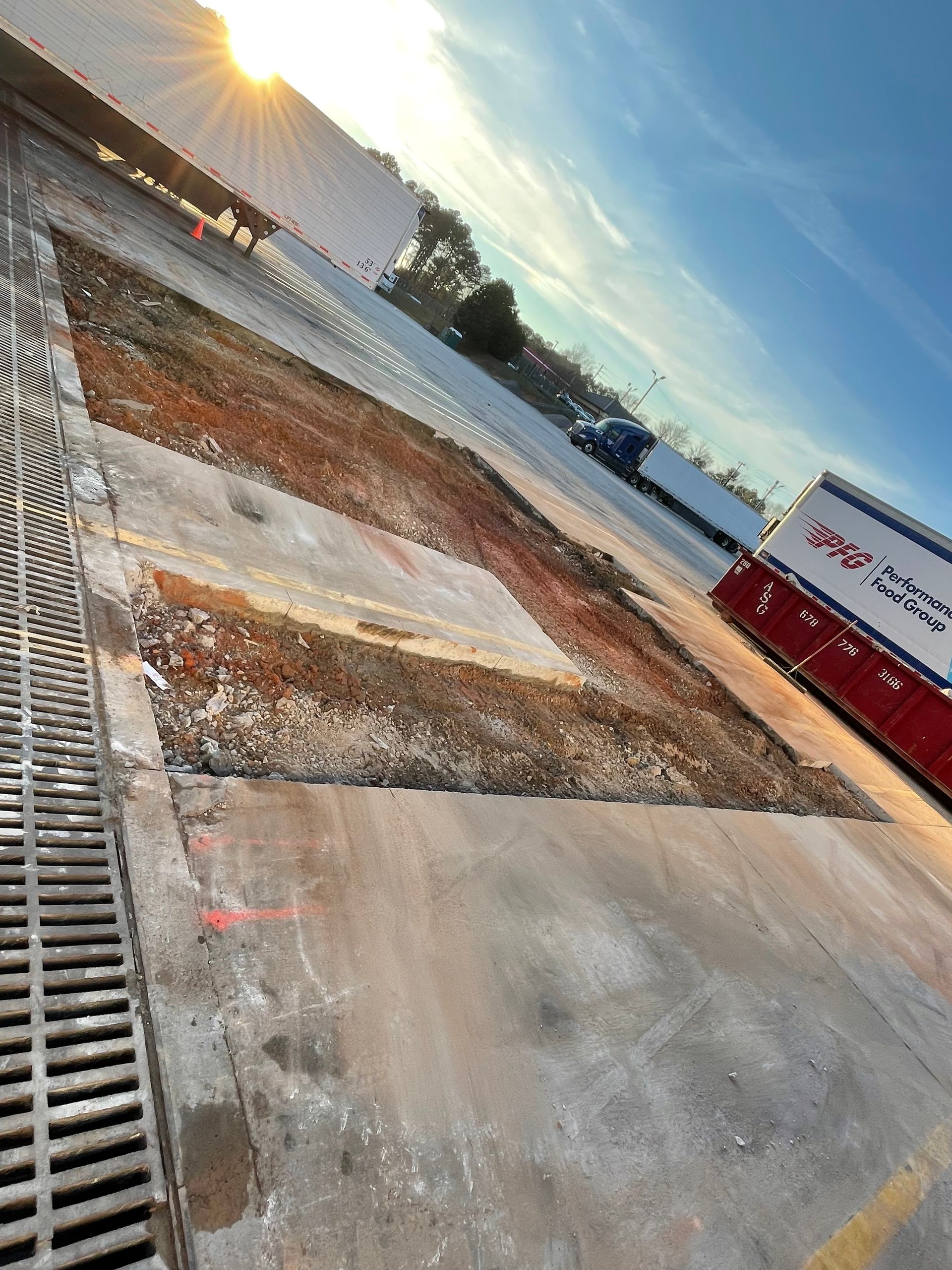 Tilted view of a construction site with a wet concrete slab, gravel, and a red-and-white trailer under blue sky