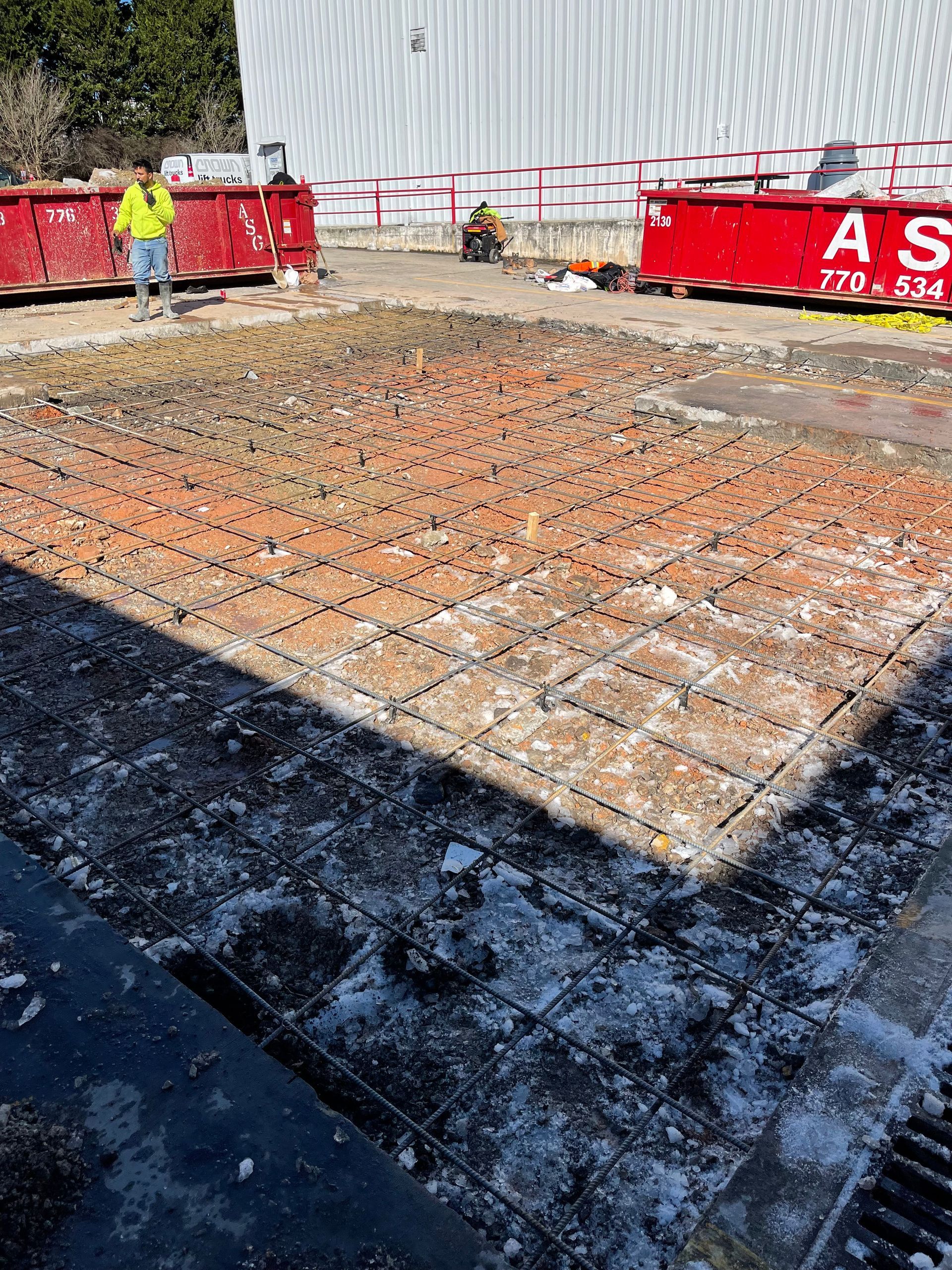 Construction site with rebar foundation slab, red barriers, and a worker in a yellow jacket.
