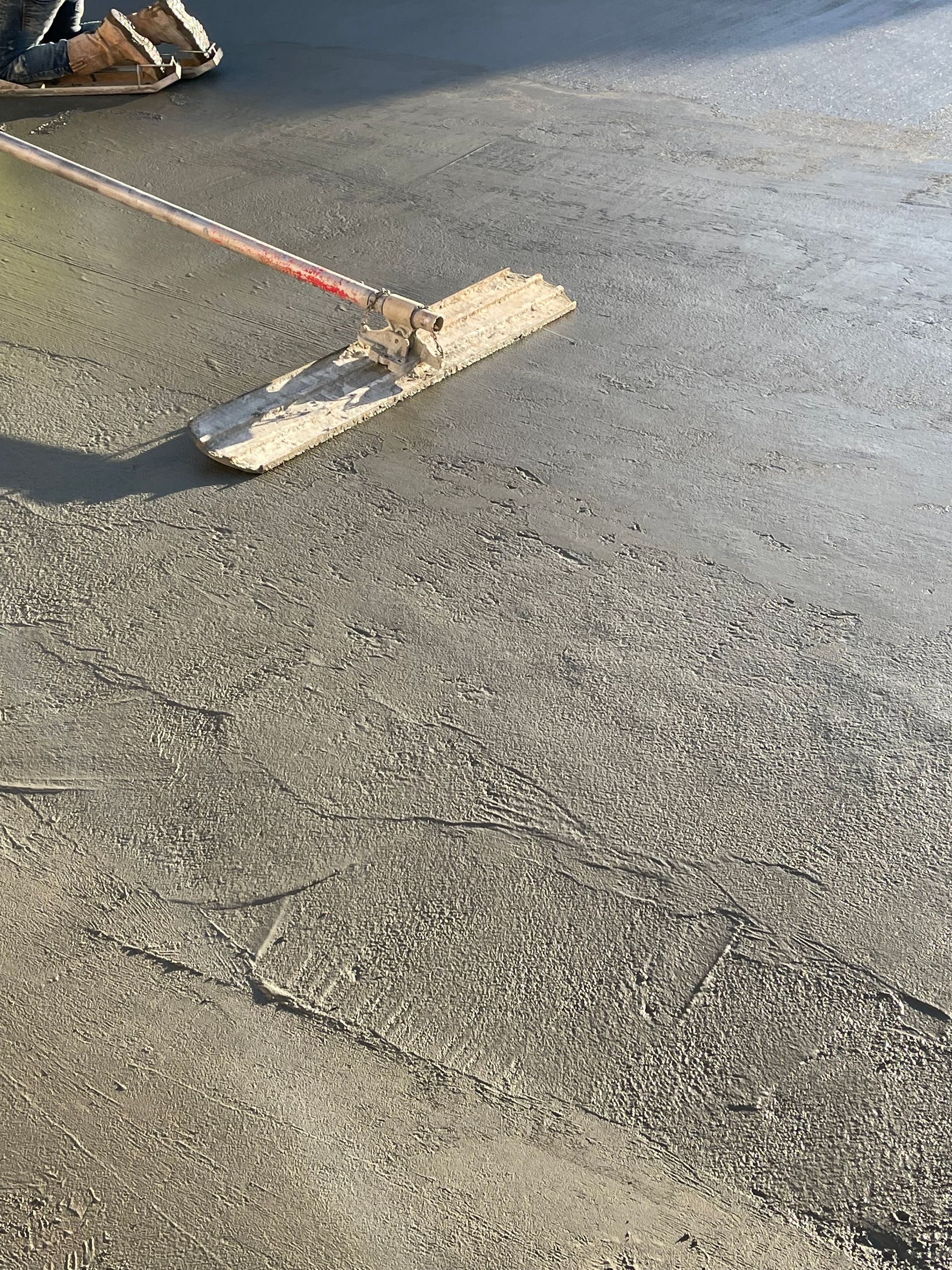 Person sweeping loose gravel off a sunlit asphalt surface with a push broom.