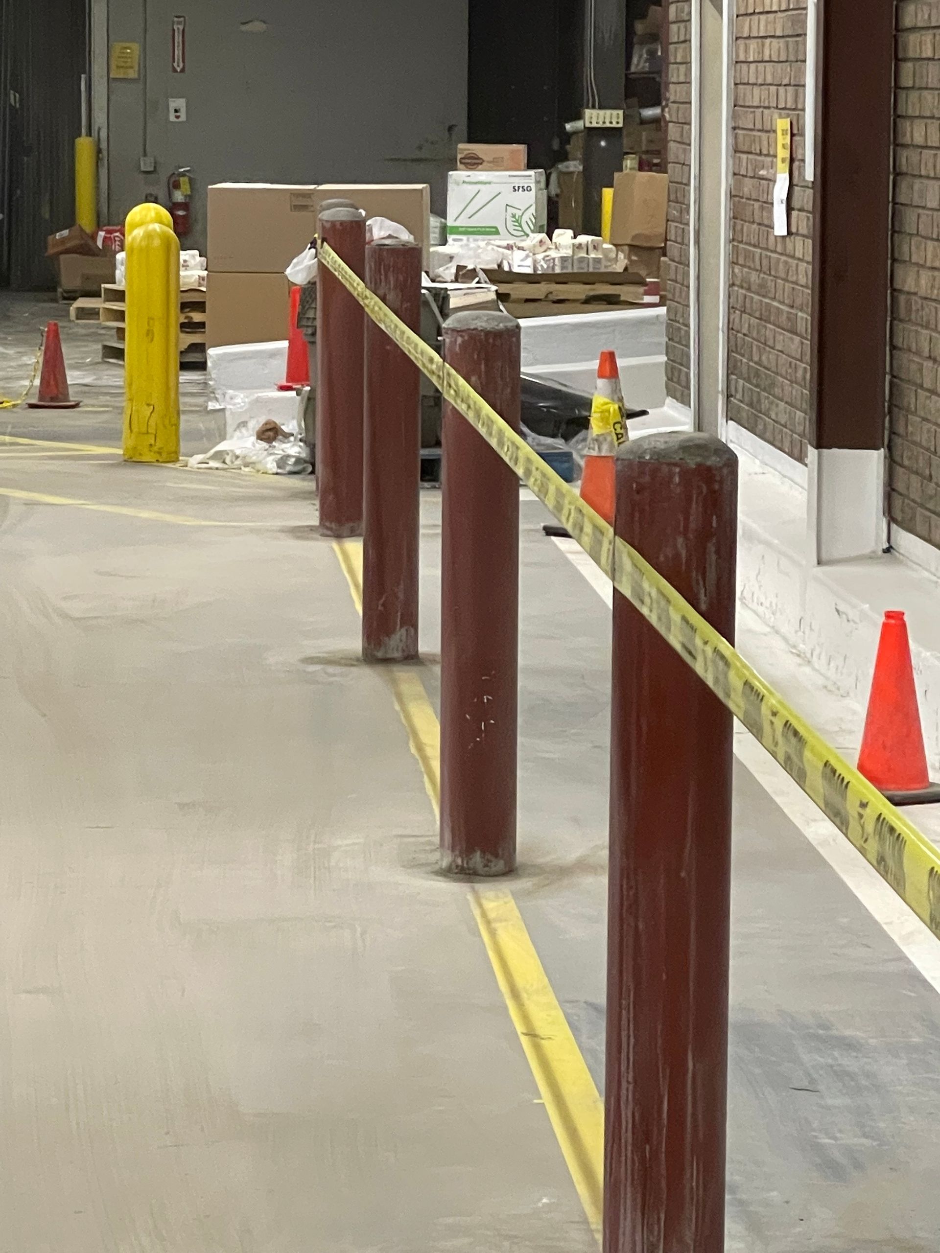 Cordoned-off sidewalk with yellow tape, orange cones, and red bollards near a building entrance.