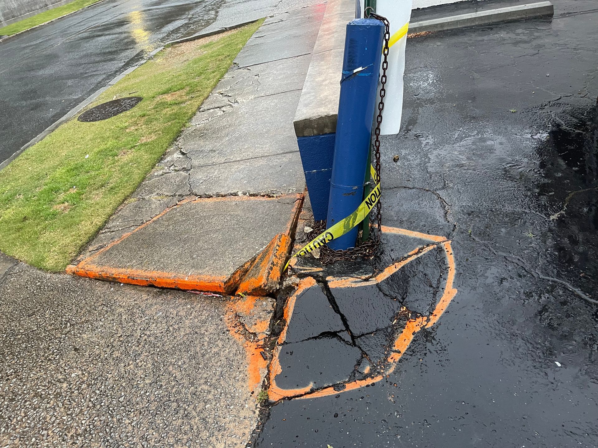 Cracked sidewalk around a blue utility pole, marked with orange paint and a broken curb near wet pavement