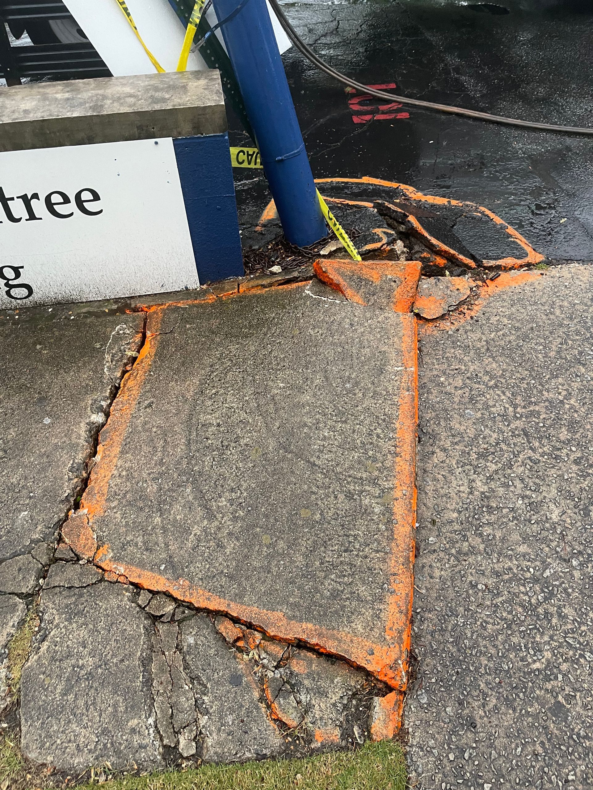 Broken sidewalk patched with orange tape near a blue pole and curbside sign