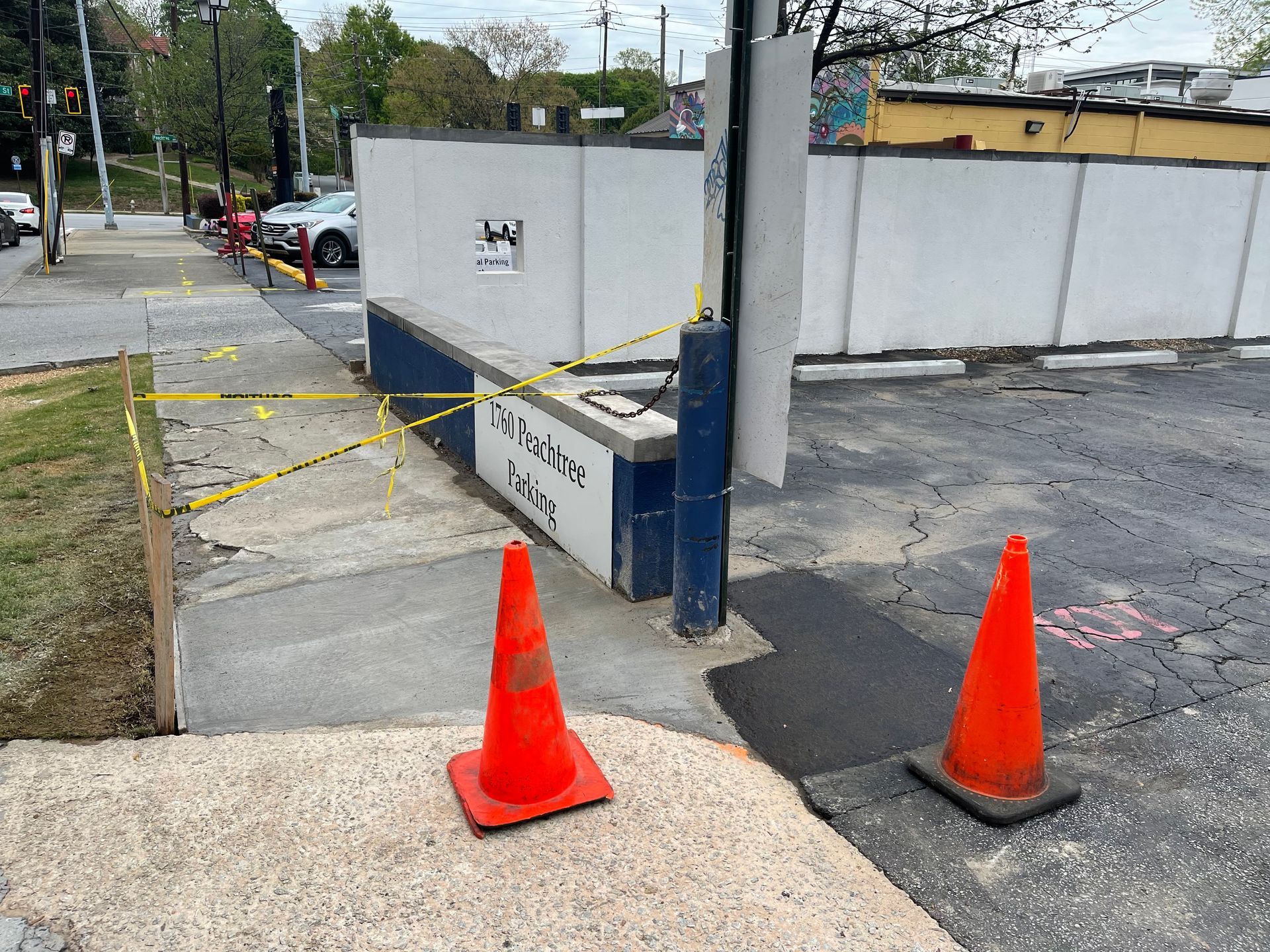 Orange cones block a sidewalk ramp beside a street, with yellow caution tape and a low wall.