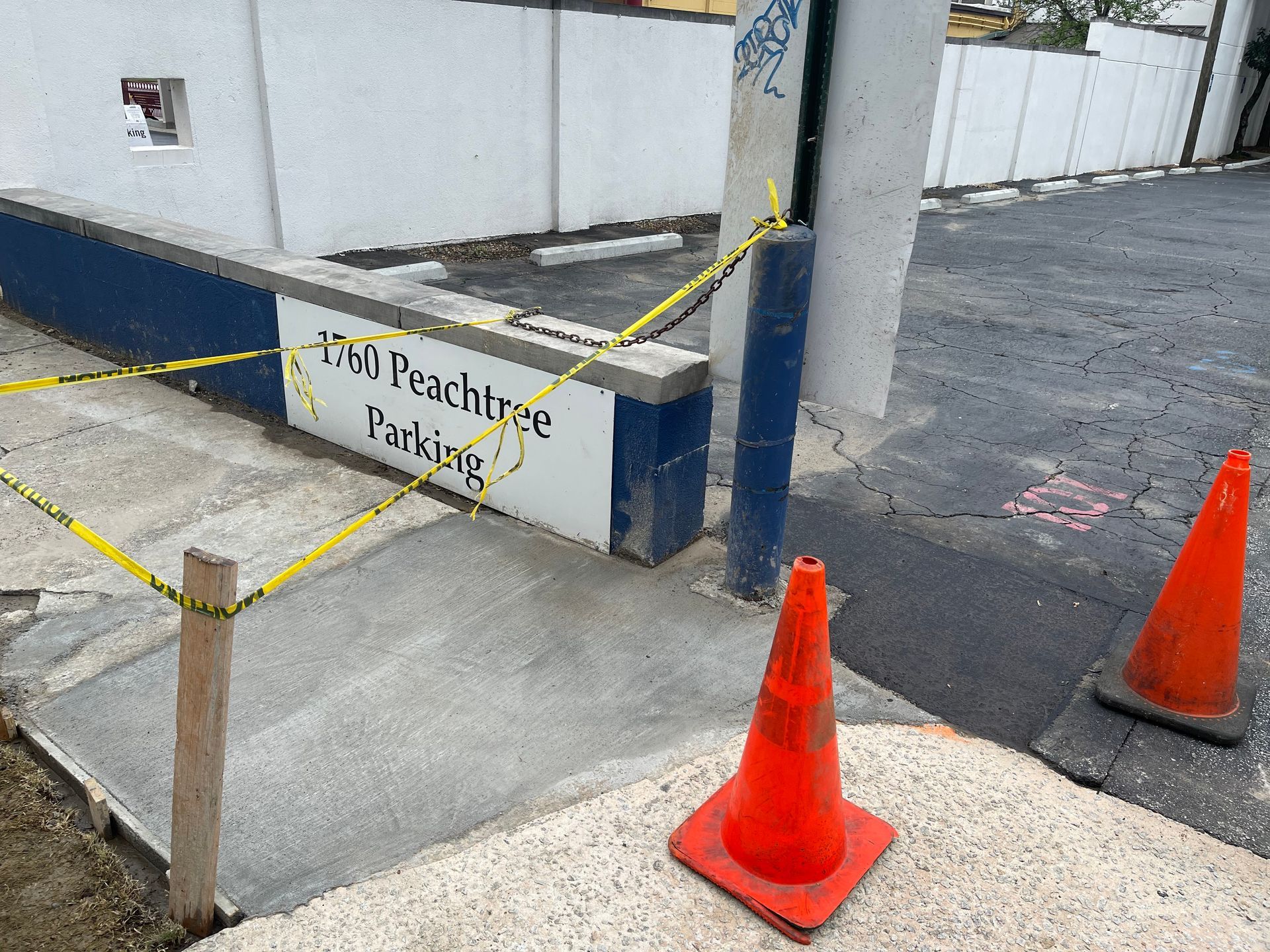 Orange cones and yellow caution tape blocking a concrete driveway beside a white wall and parking area