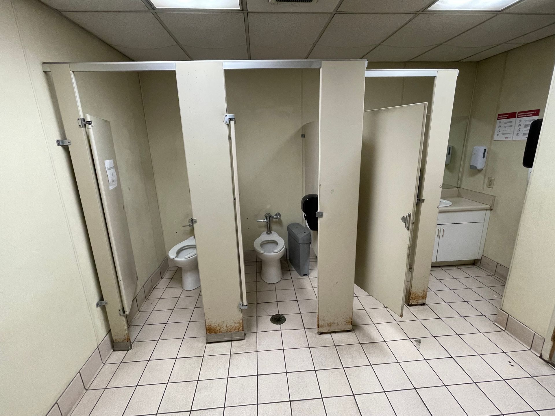 Public restroom with three toilet stalls and a tiled floor, one stall showing a toilet and one open doorway
