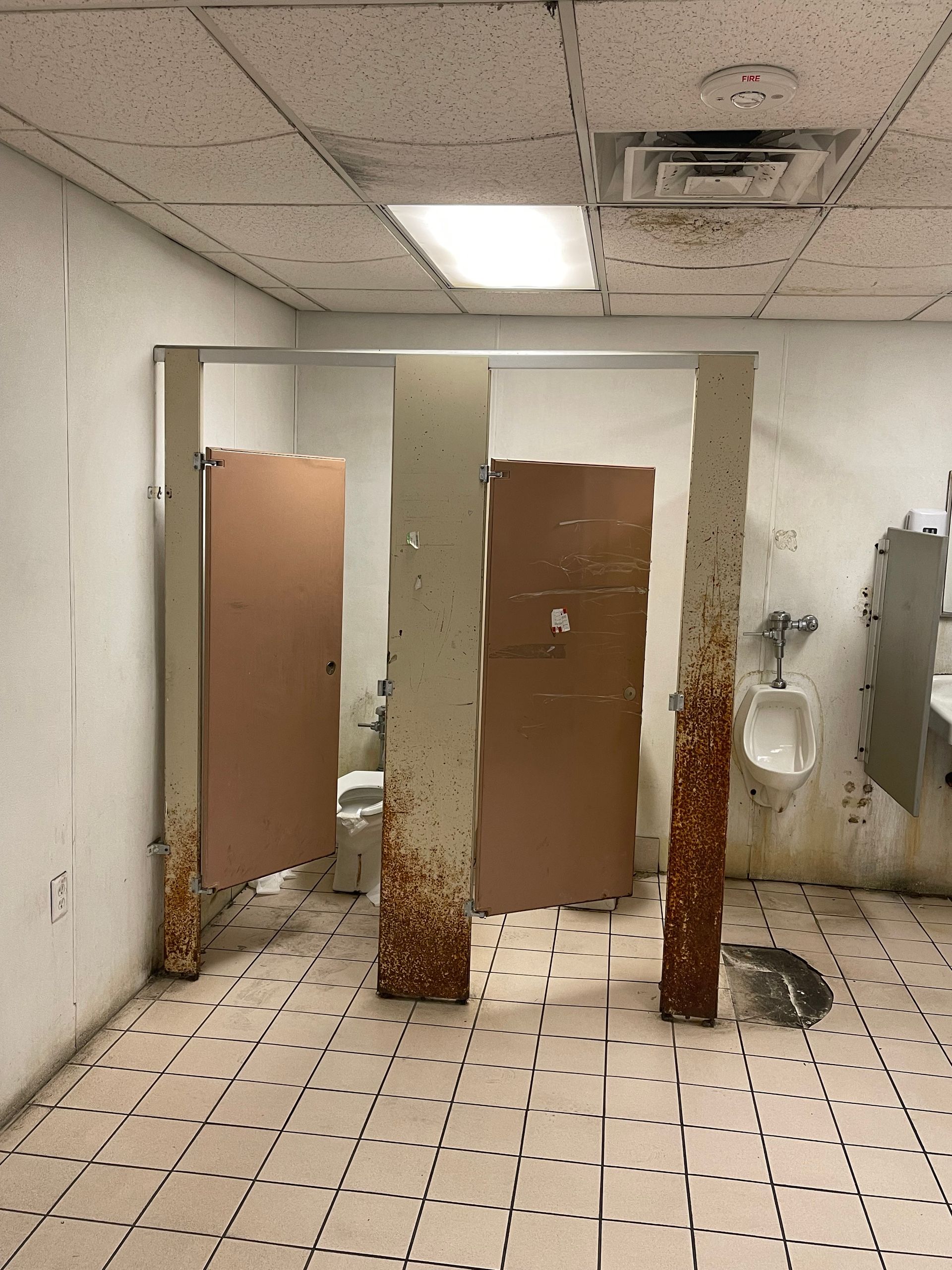 Run-down restroom with rust-stained stall dividers, tiled floor, and a urinal on the right.
