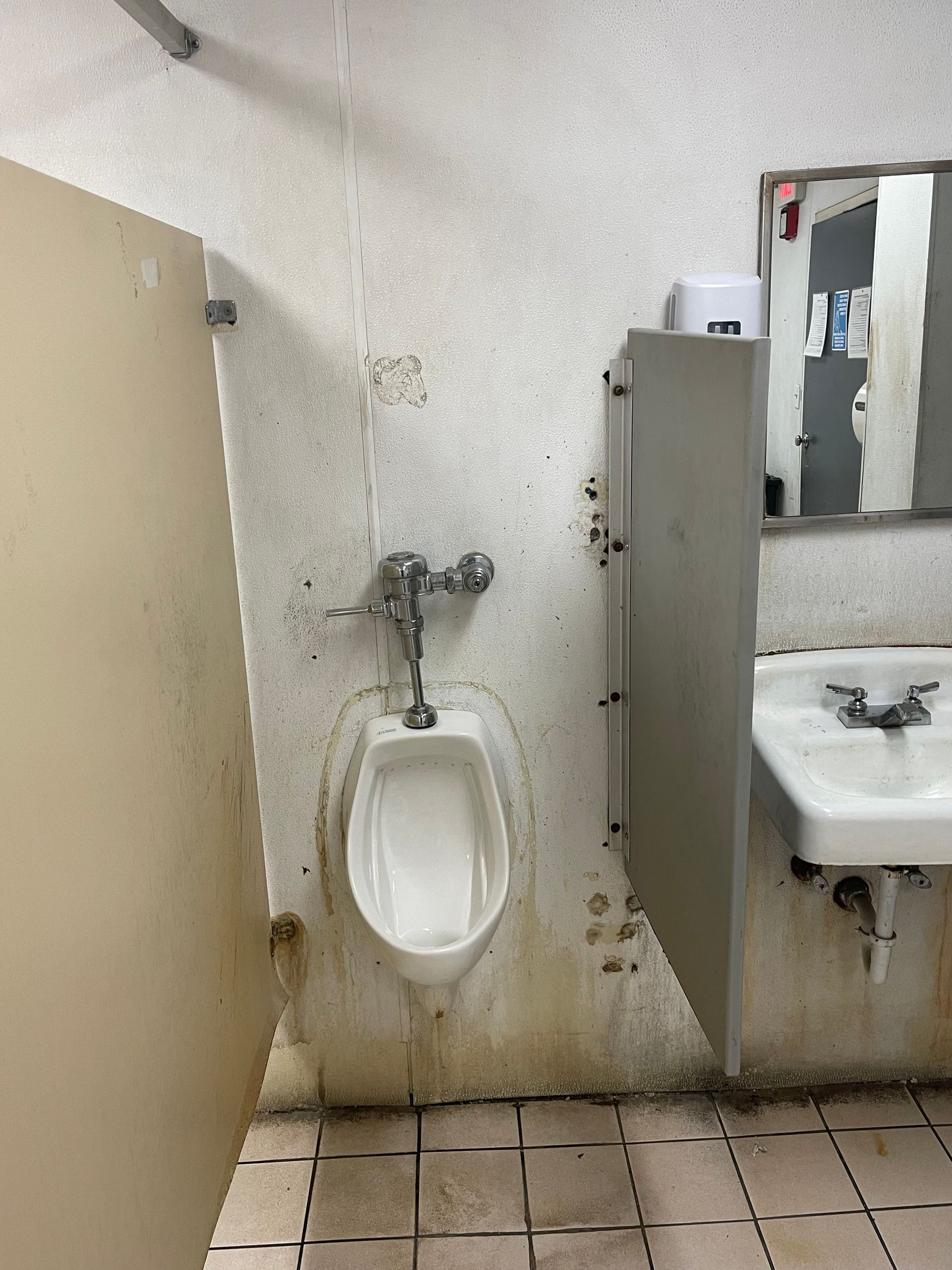 Public restroom with a urinal between beige stalls and a sink on the right; wall and floor are stained.