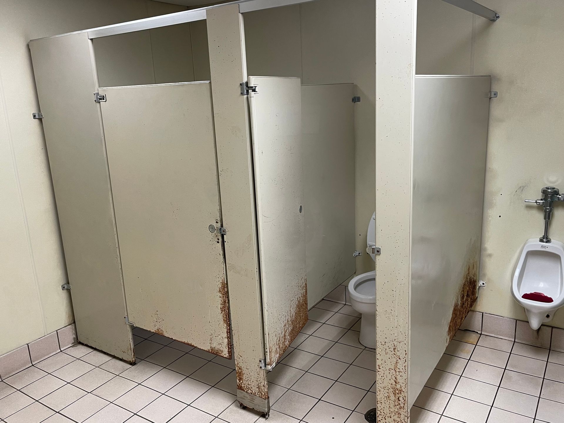 Public restroom stalls with beige dividers and a wall-mounted urinal, showing visible rust and grime.
