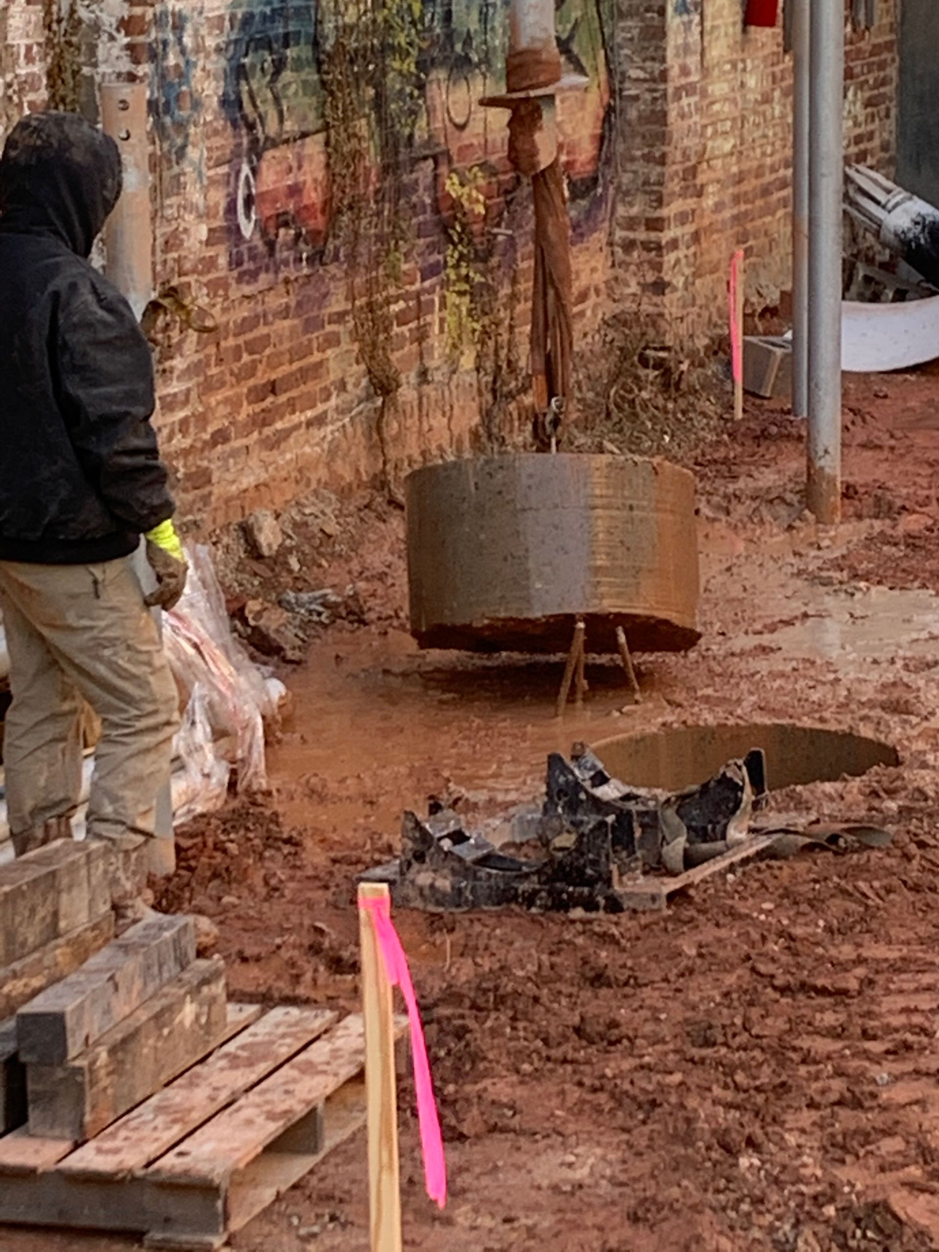 Construction worker beside excavator digging near a brick wall and exposed soil, with a burned debris pile in foreground