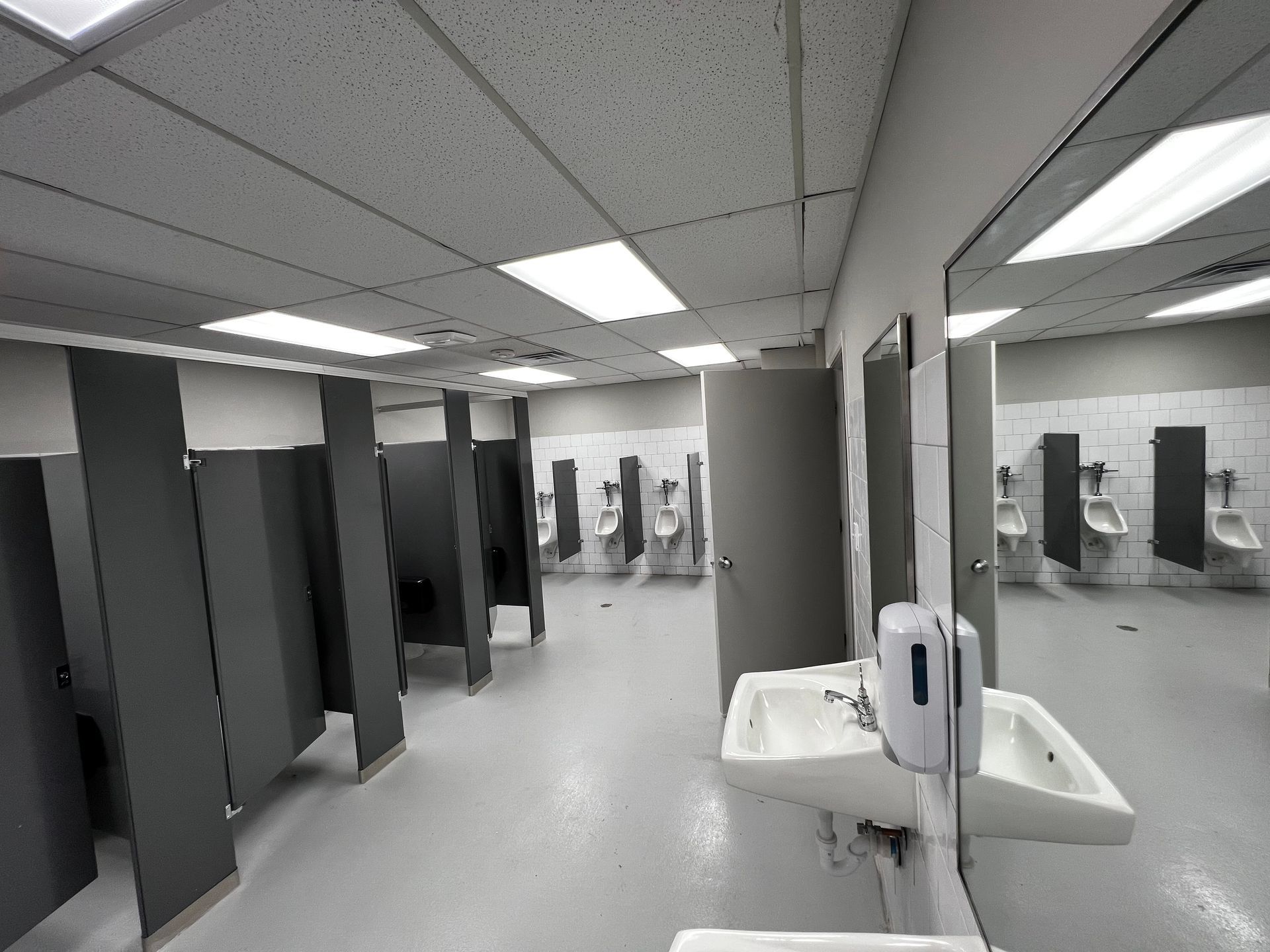 Public restroom with gray stalls and sinks under fluorescent lighting, viewed from inside near the mirrors.
