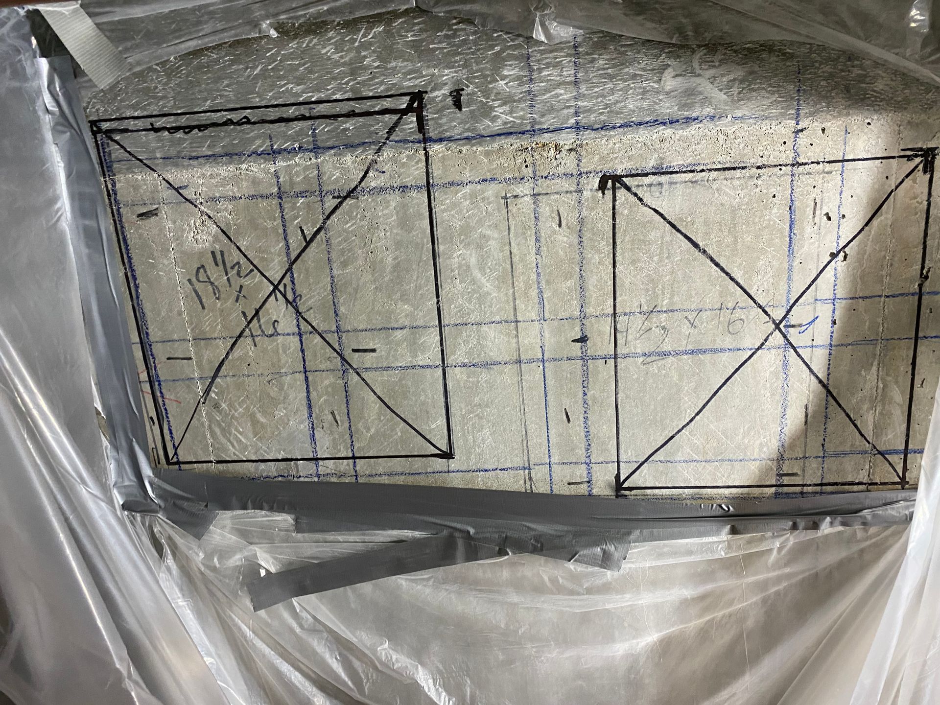 Two framed window screens wrapped in plastic, with black tape forming X shapes on each panel.