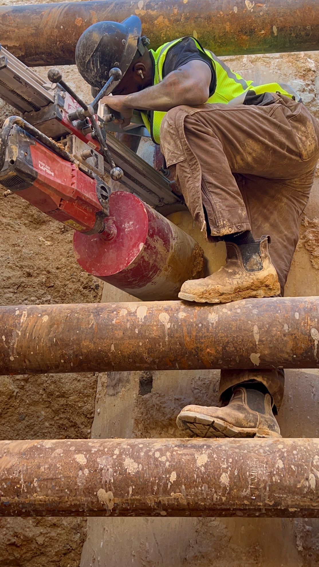 Worker in a hard hat uses a red power tool while crouched among large rusted pipes.