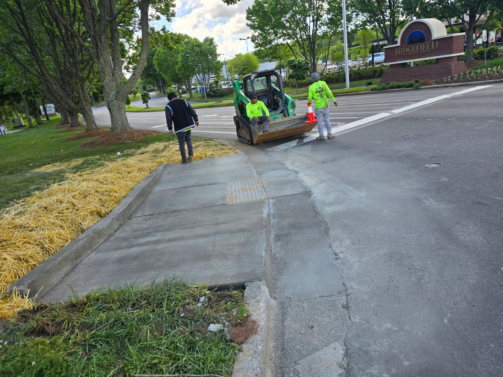 Workers repairing a roadside curb and sidewalk beside a street, with a skid steer and fresh concrete.