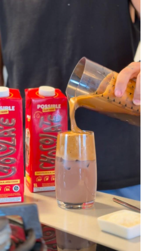 Uma mão despeja espuma de café em um copo de leite com chocolate e cubos de gelo. Duas caixas vermelhas com a palavra