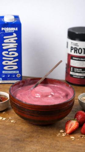 Tigela de smoothie rosa com colher, rodeada de ingredientes como morangos e proteína em pó.