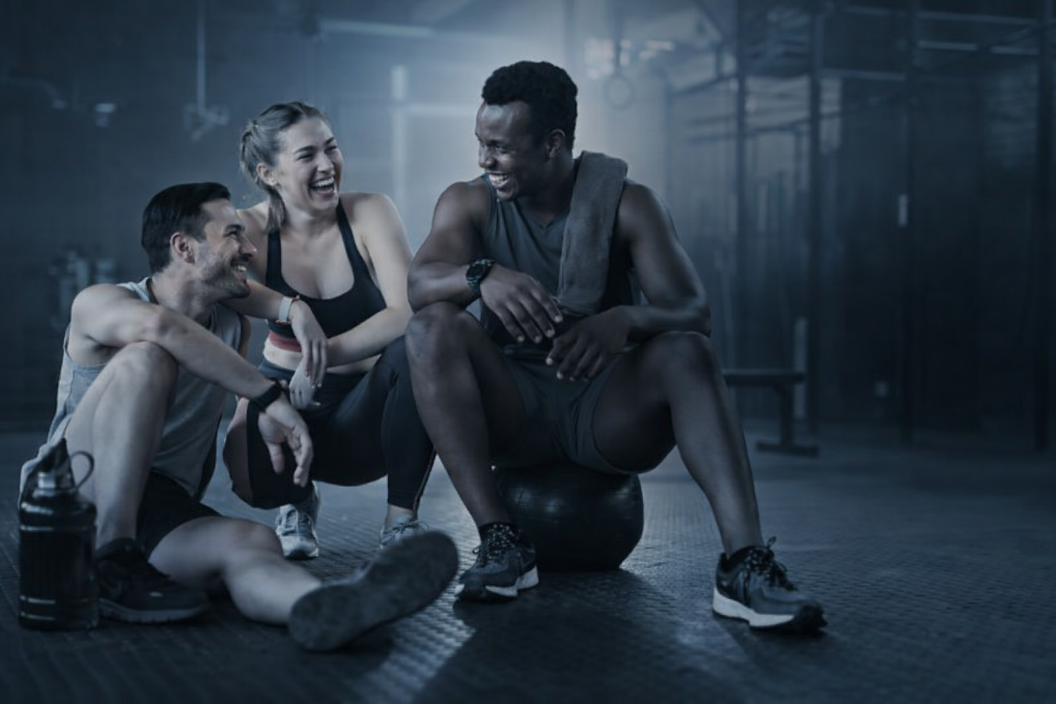Três pessoas rindo juntas em uma academia. Uma mulher e dois homens relaxam e sorriem após o treino.