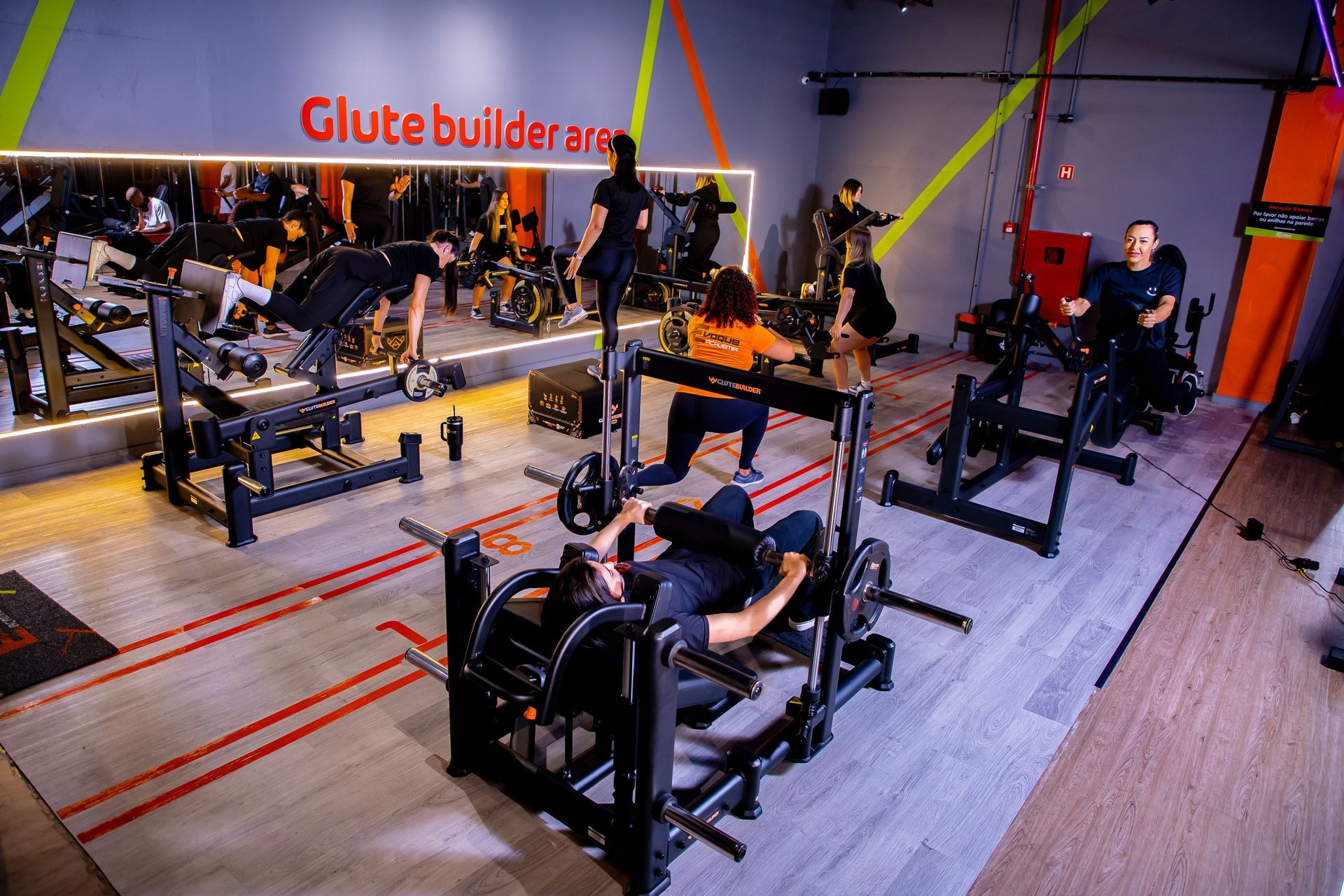 Interior de academia com pessoas usando aparelhos de musculação. Detalhes em vermelho, laranja e amarelo nas paredes e nos equipamentos.