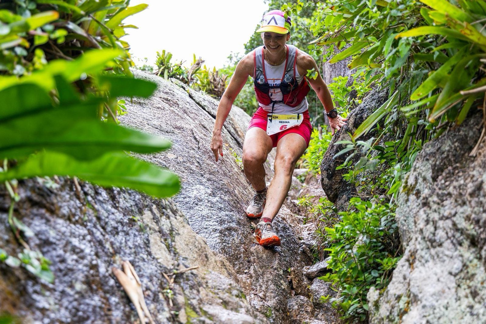 Corredor de trilha desce uma encosta rochosa em uma floresta. Vestindo shorts vermelhos, colete de hidratação e boné. Sorrindo.