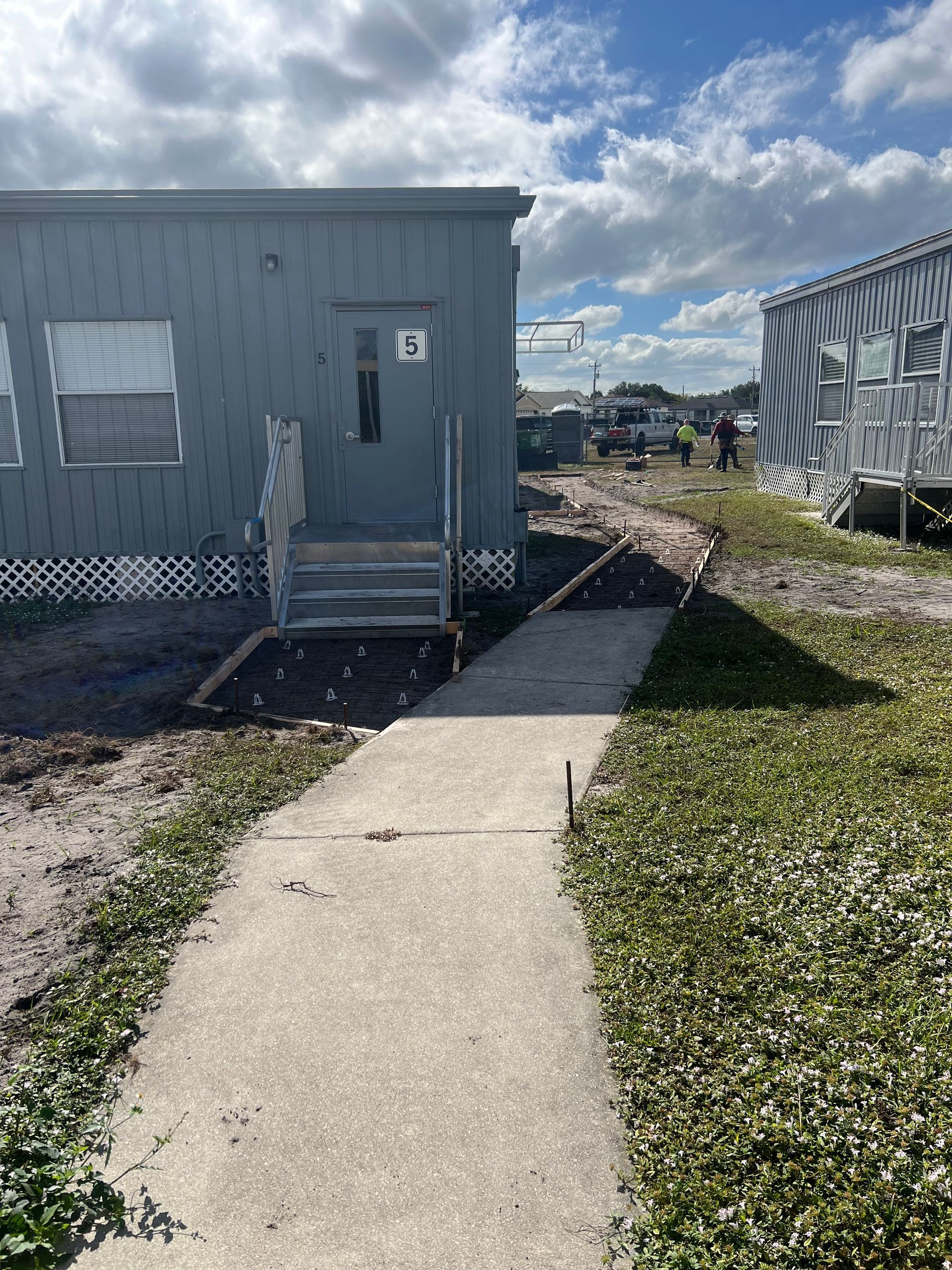 A concrete walkway leads to a gray modular building with steps. Another building is to the right.