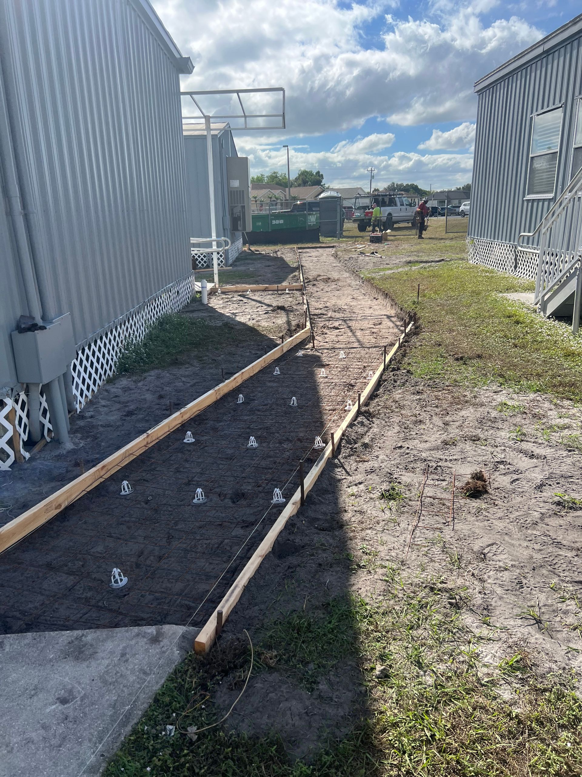 A narrow dirt path under construction between two buildings, bordered by wooden beams and dark soil, on a sunny day.
