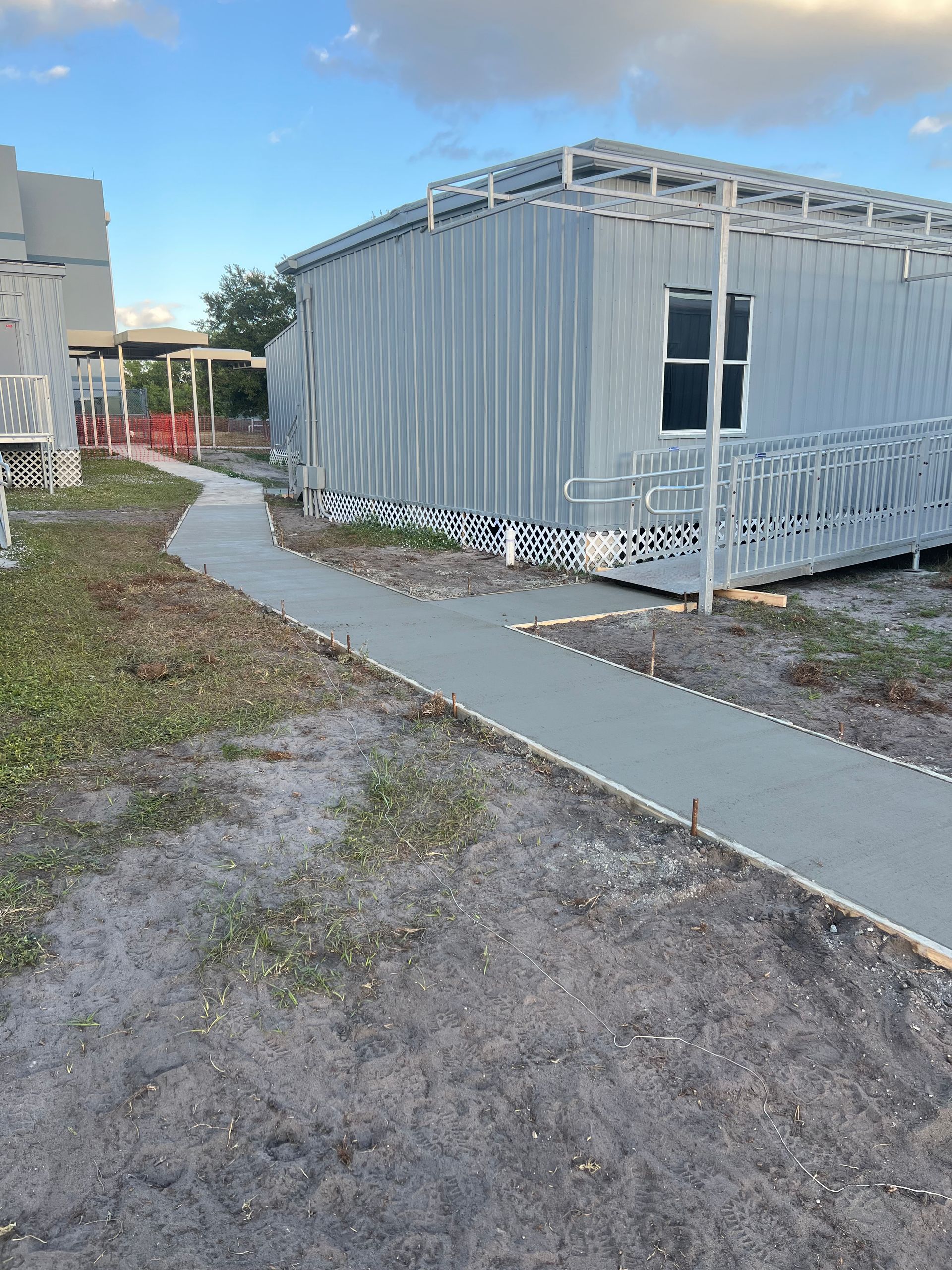 Newly poured concrete pathway leads to a prefabricated metal building with a ramp, under a cloudy sky.