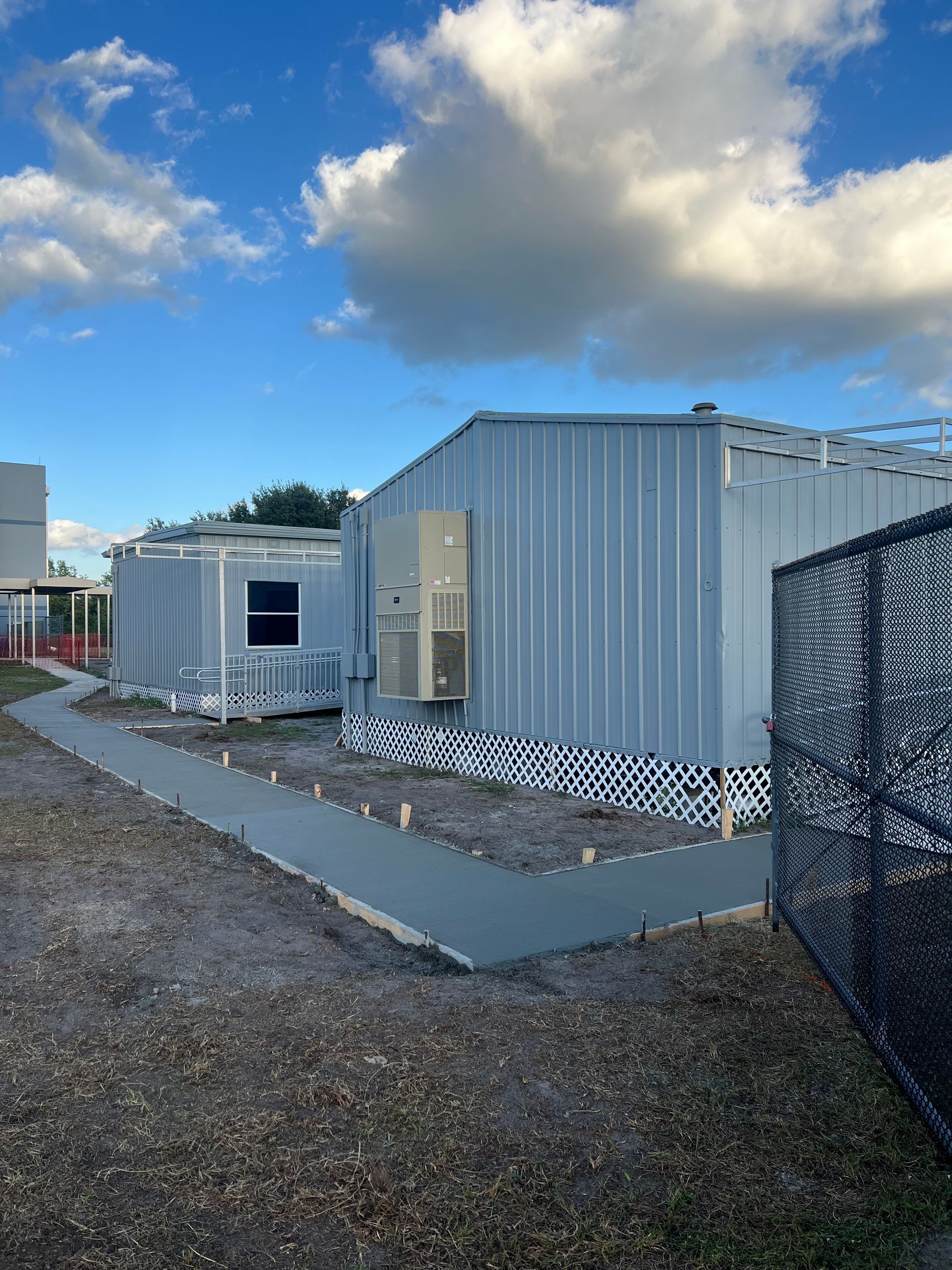 Modular building with a concrete walkway, air conditioner, and chain-link fence on a cloudy day.