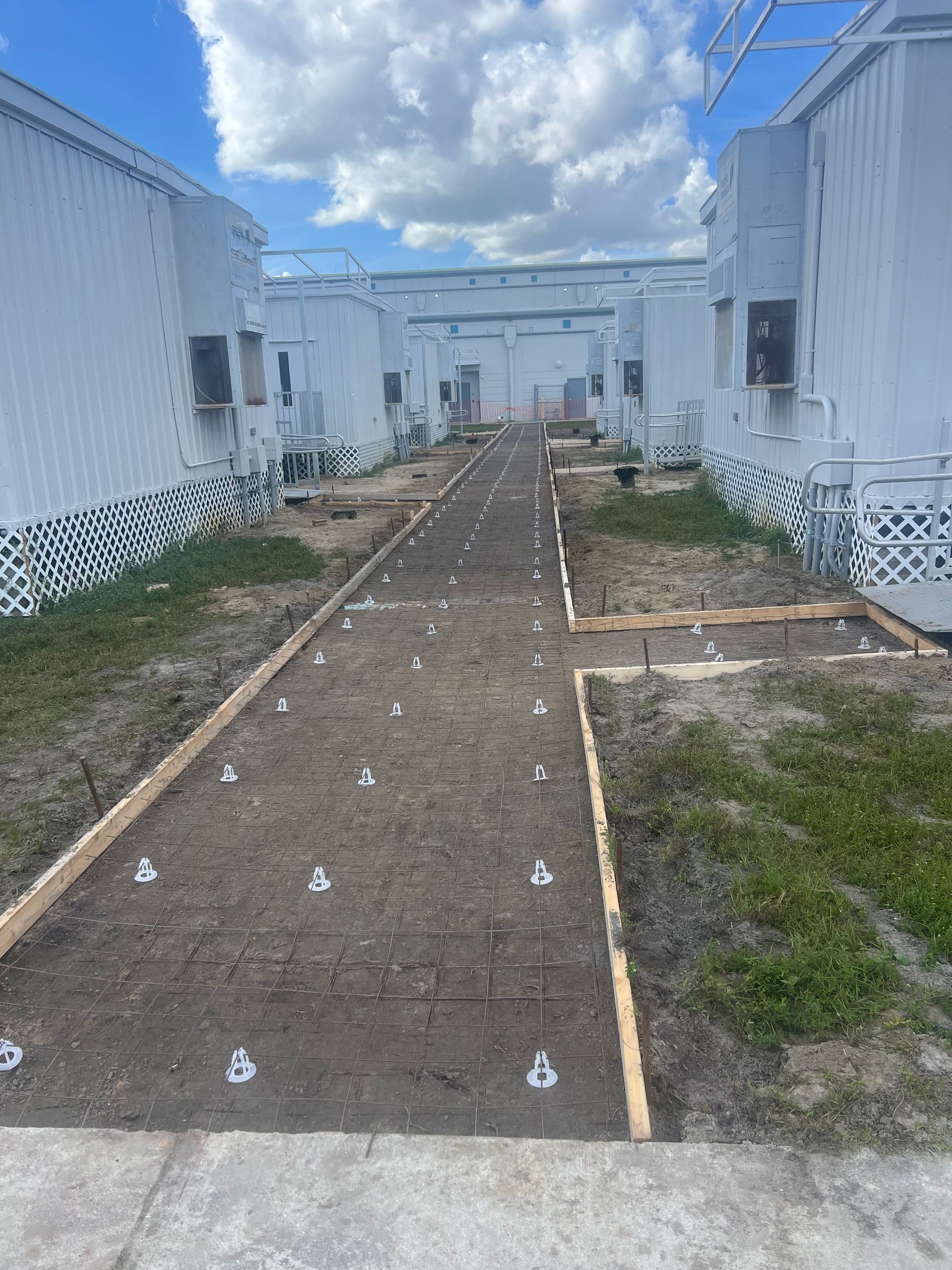 Pathway between white modular buildings, dirt and concrete with anchors. Blue sky.