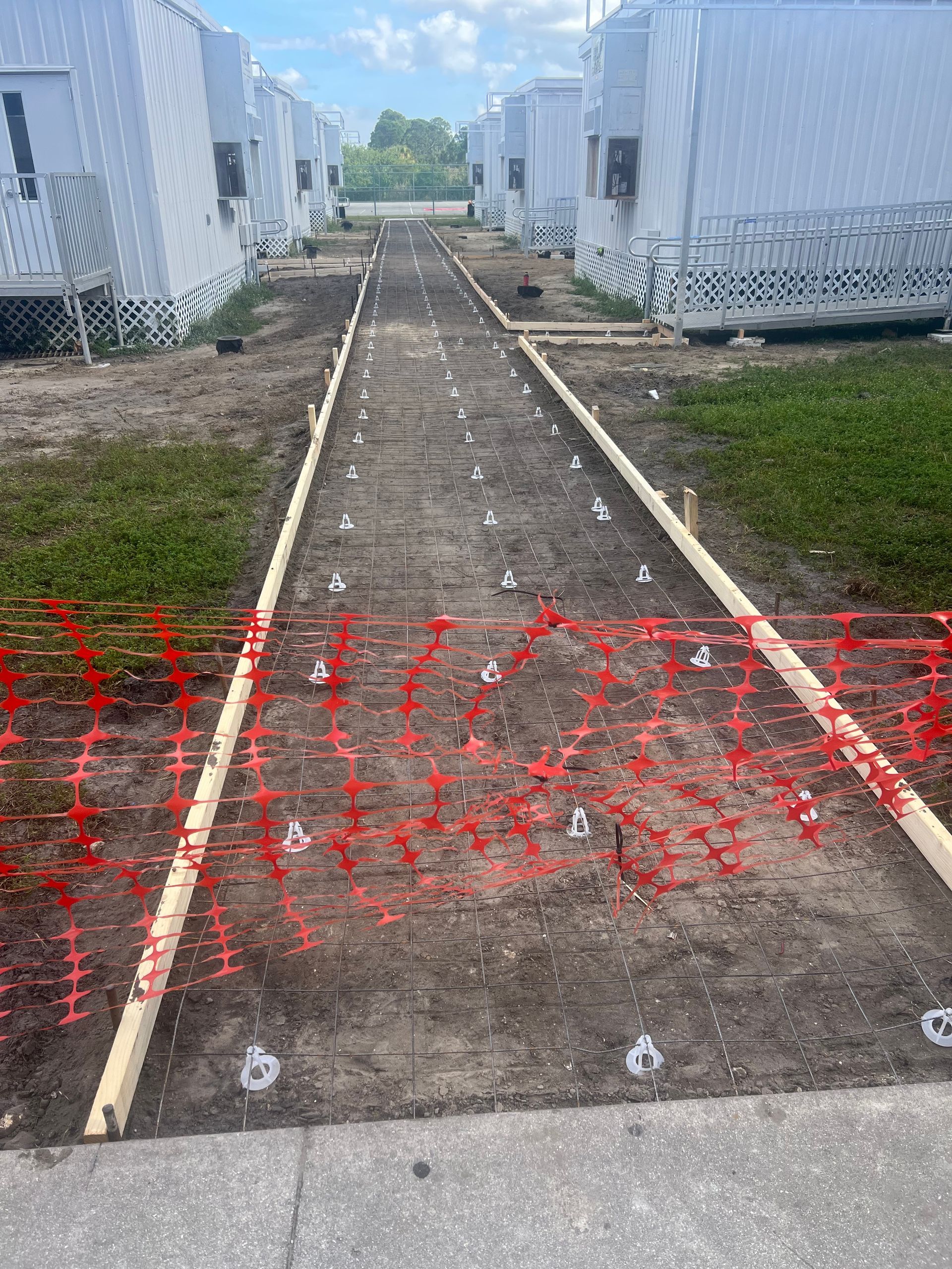 A construction site with wooden forms and orange fencing; preparing to pour concrete walkway between white modular buildings.