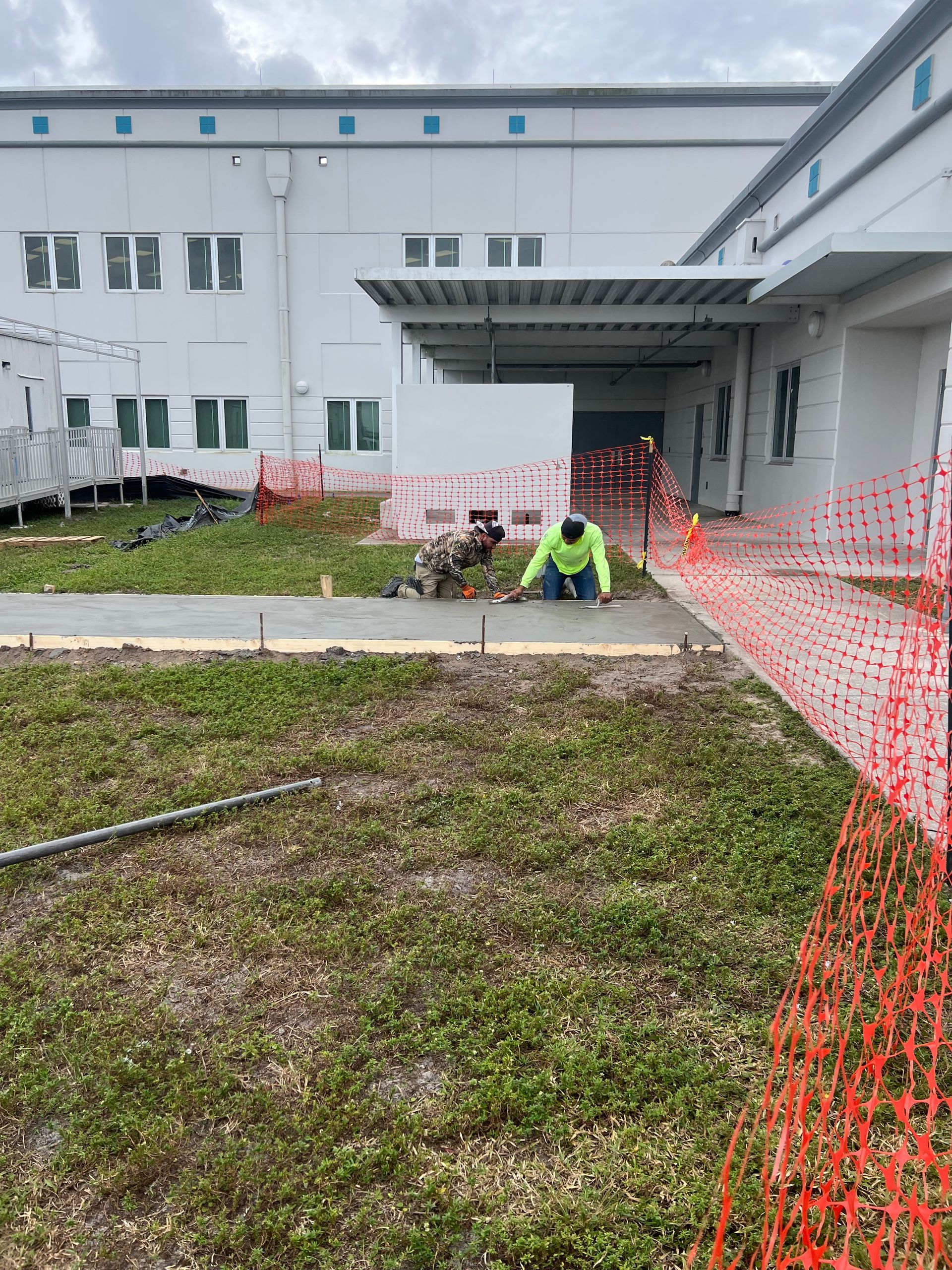 Construction worker kneeling, smoothing concrete sidewalk. Exterior shot with building in background, orange safety fence.
