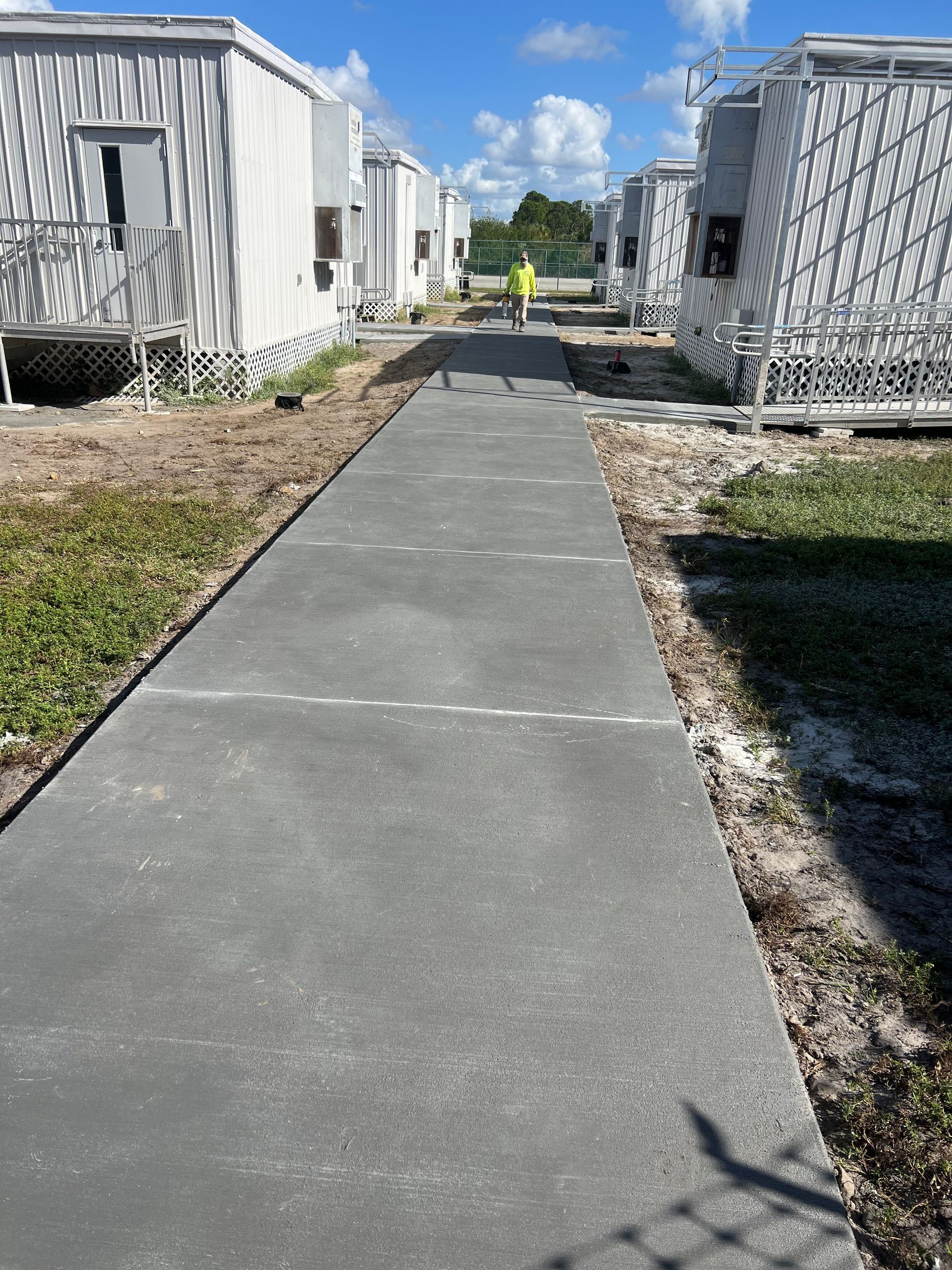 Concrete walkway between gray modular buildings. A person in a high-vis vest walks away from the camera.