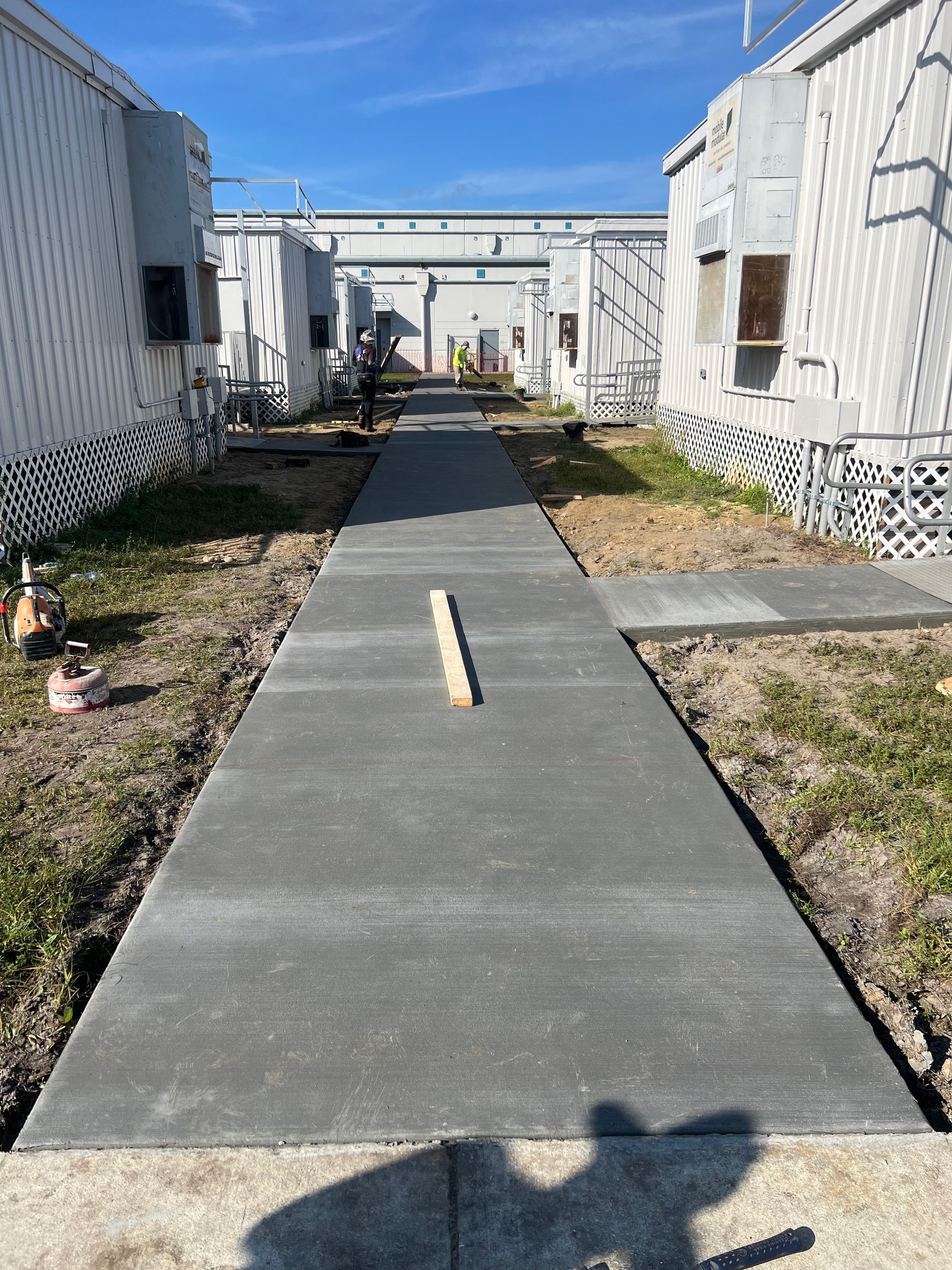 Concrete walkway between modular buildings under a blue sky; construction in progress.