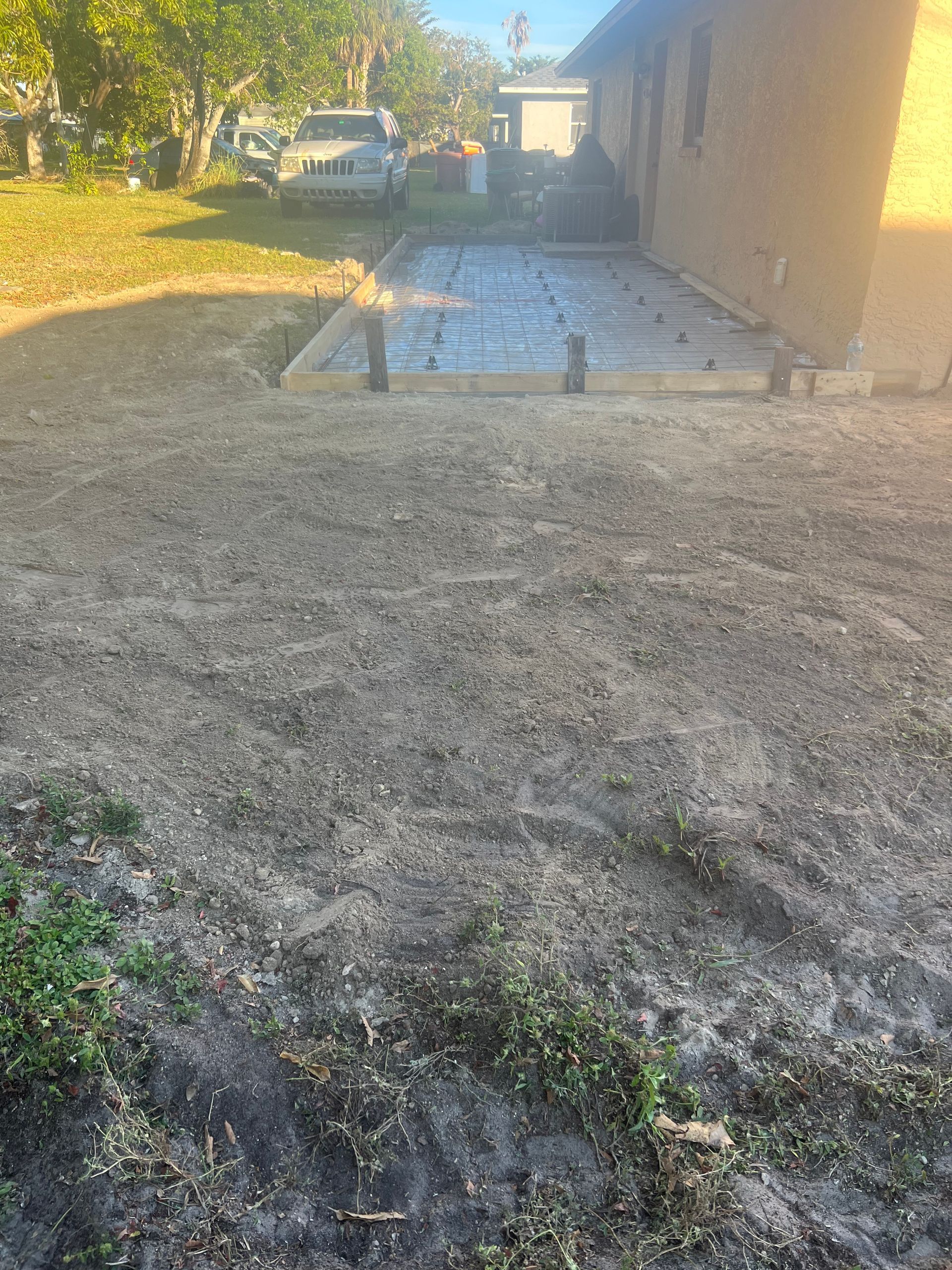 Backyard with newly poured concrete patio, surrounded by dirt and grass. A light-colored house is on the right.