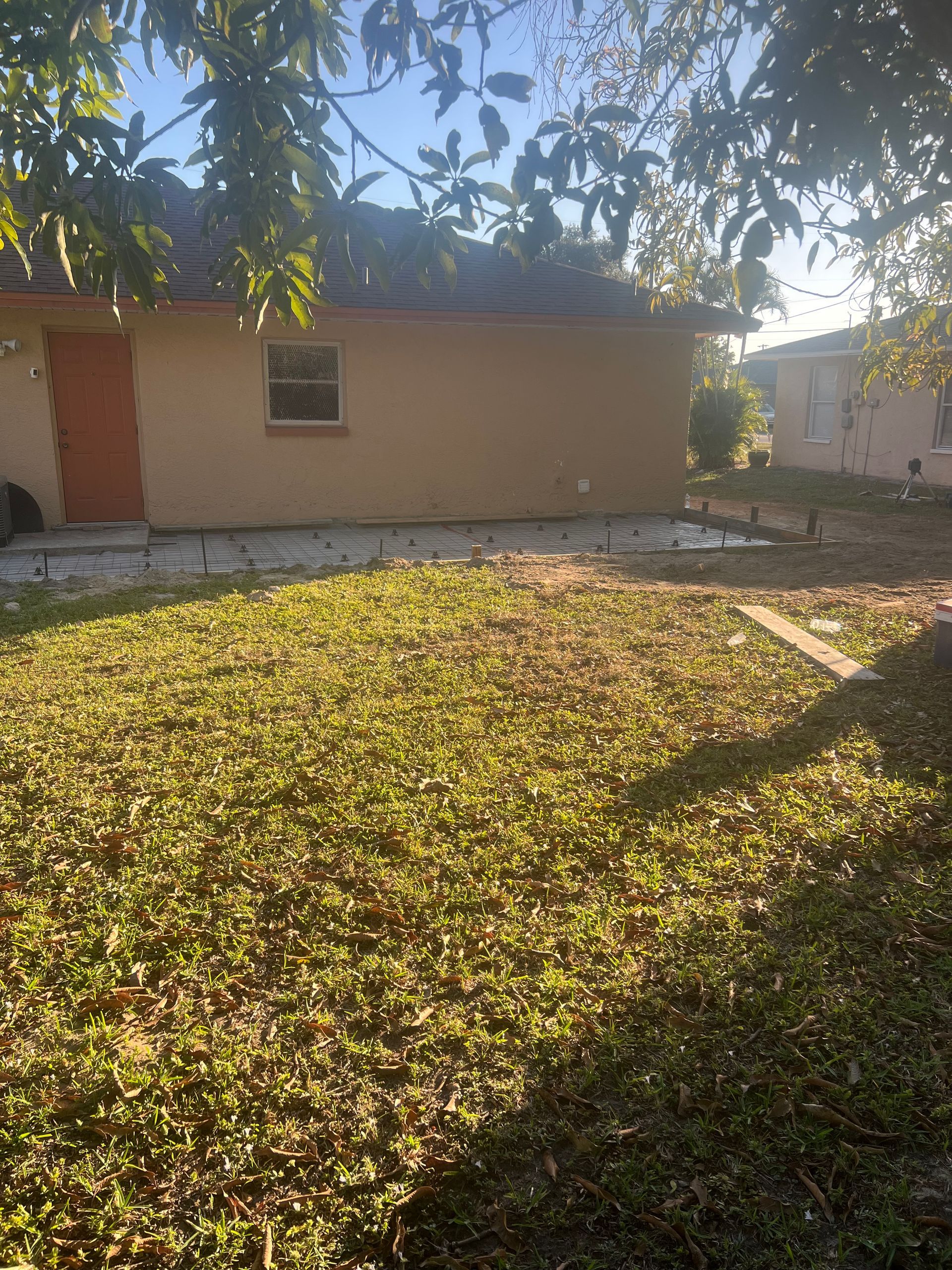 A tan house with an orange door and small window sits on a grassy yard, under tree branches.